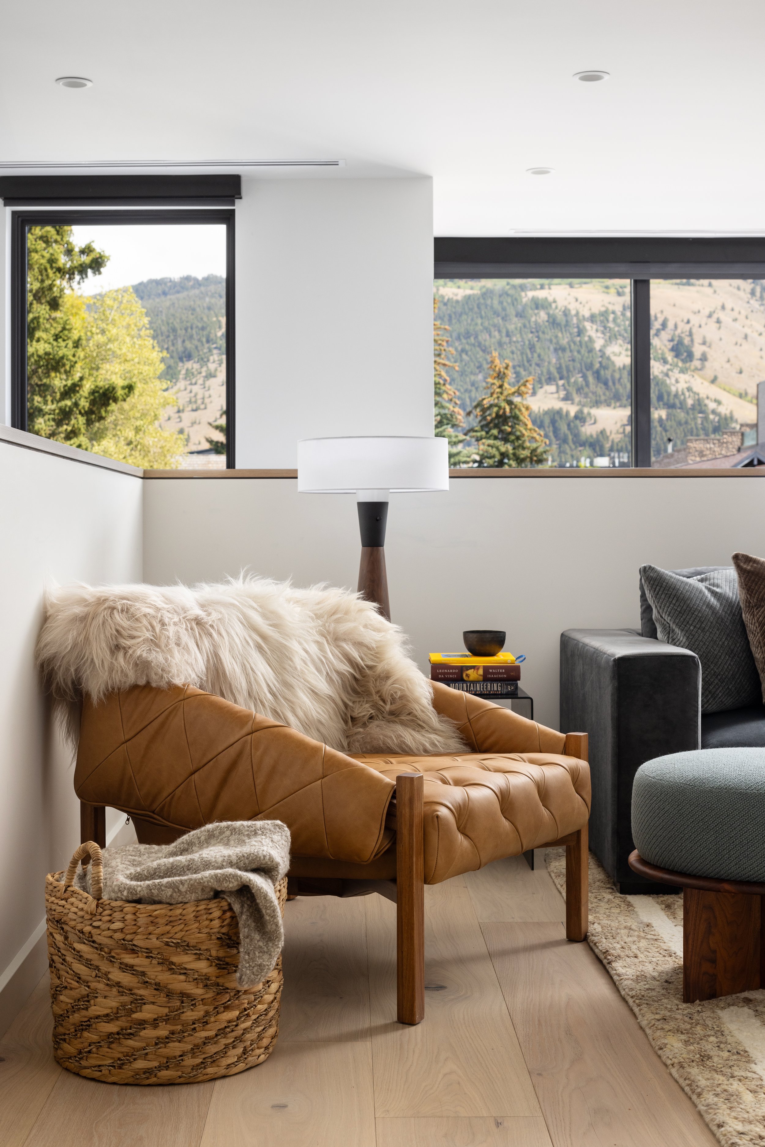 Living room with a tan leather chair, white fur throw, woven basket, black side table with books, lamps, window view of trees and mountains.