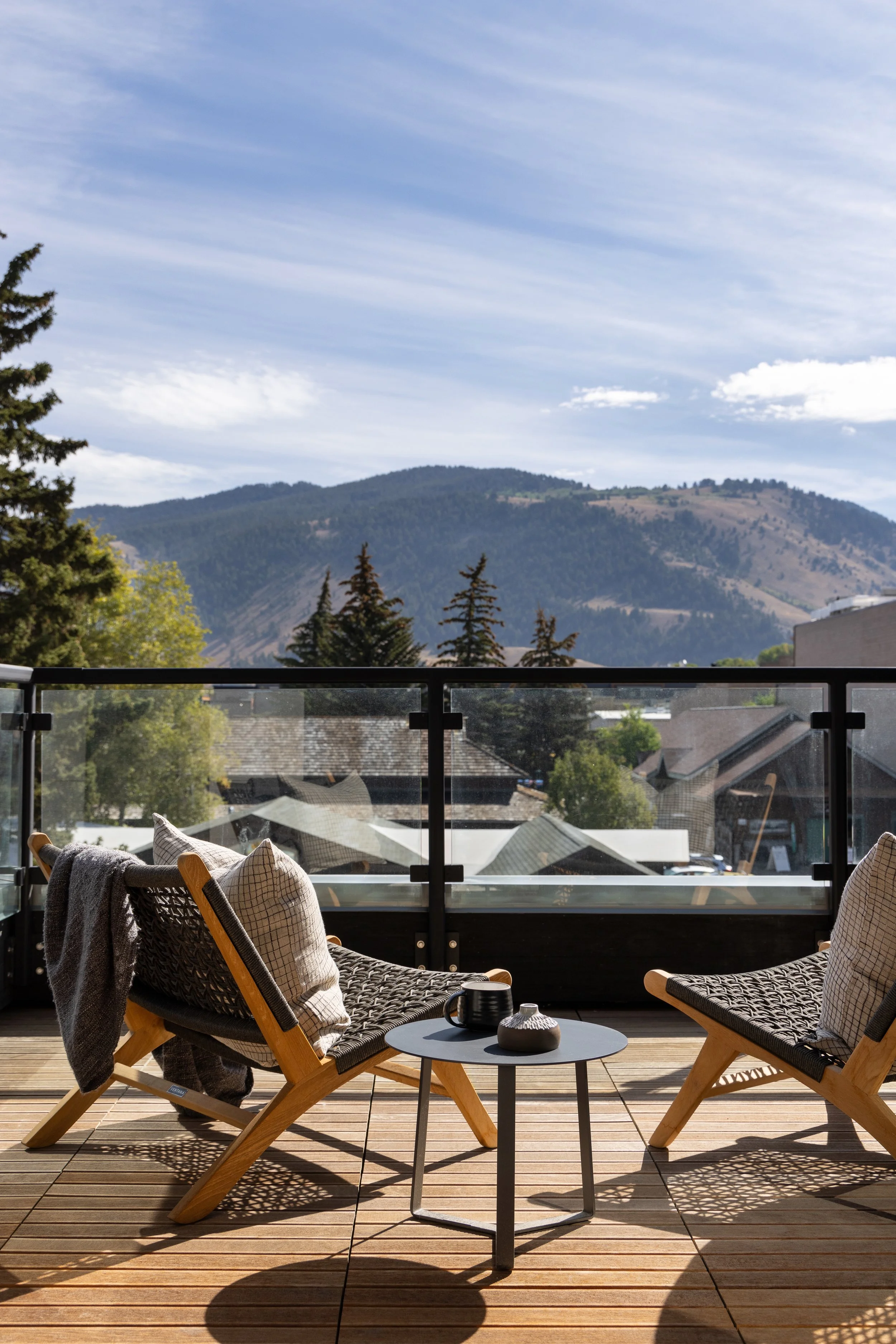 Balcony with two wooden chairs, a small round table with black cups and a decorative vase, overlooking a mountain range with trees and a cloudy sky.