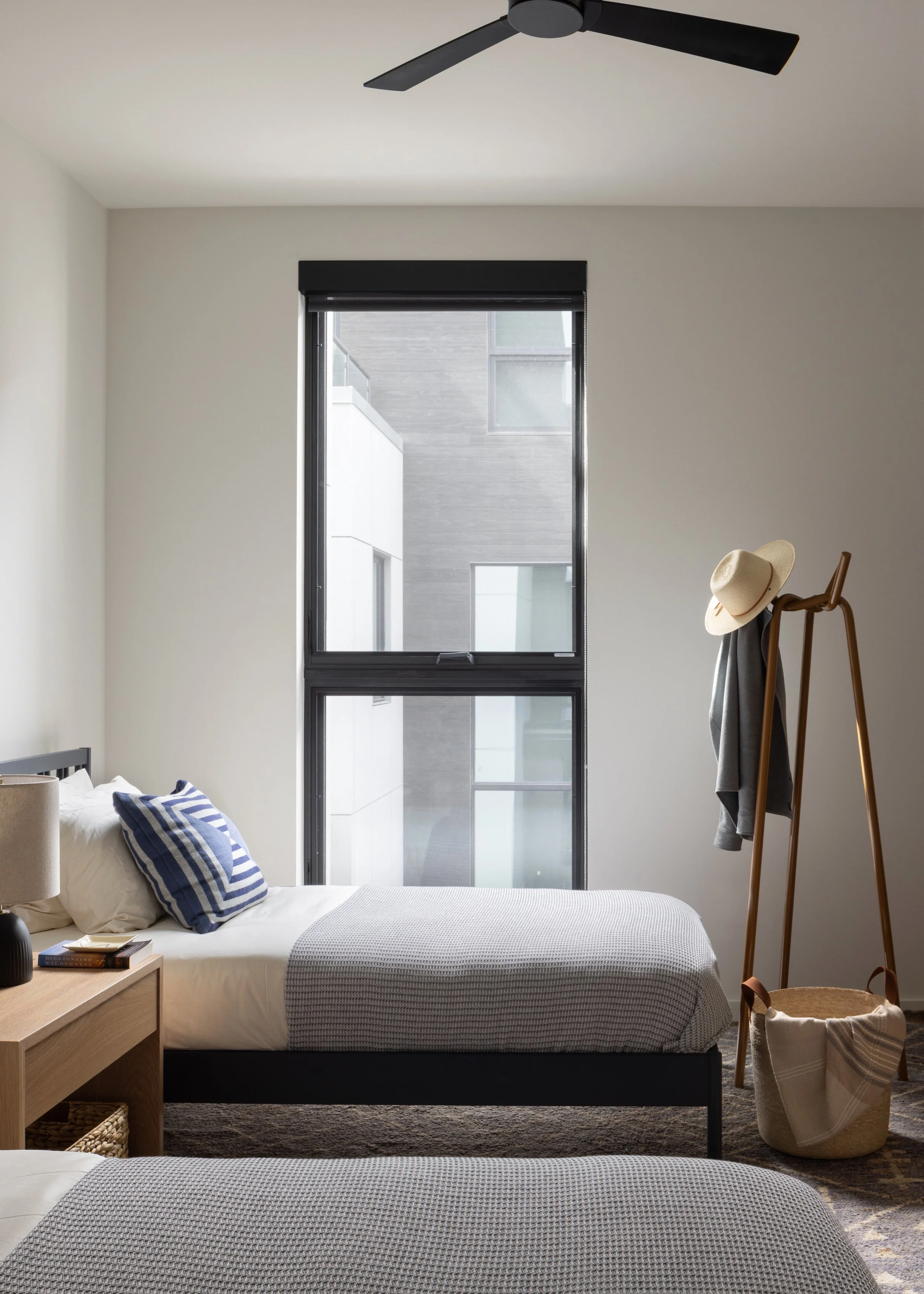 A bedroom with two twin beds, each with white bedding and a blue-striped pillow. There is a nightstand with a lamp and books between the beds. A large window is centered on the wall, with a view of a neighboring modern building. A wooden coat rack wi