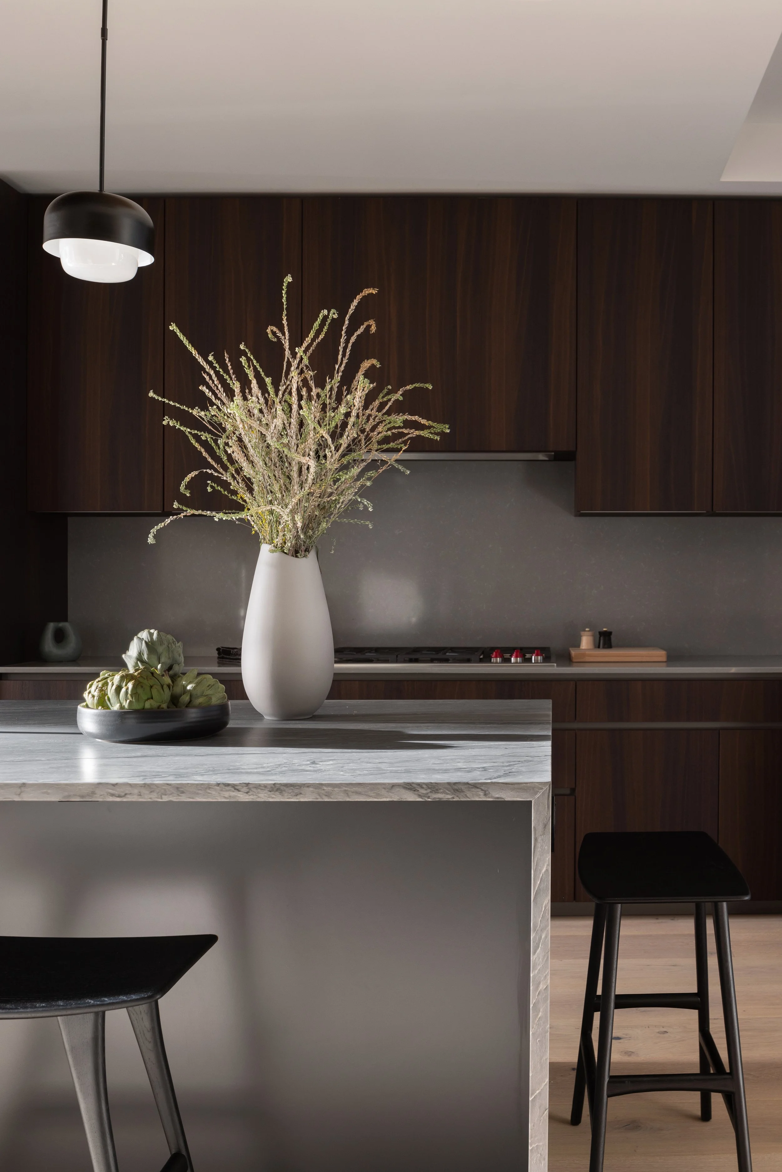 Modern kitchen with dark wood cabinets, marble countertop, and a large white vase with dried plants on the island. There is a black bowl with artichokes and a black stool.