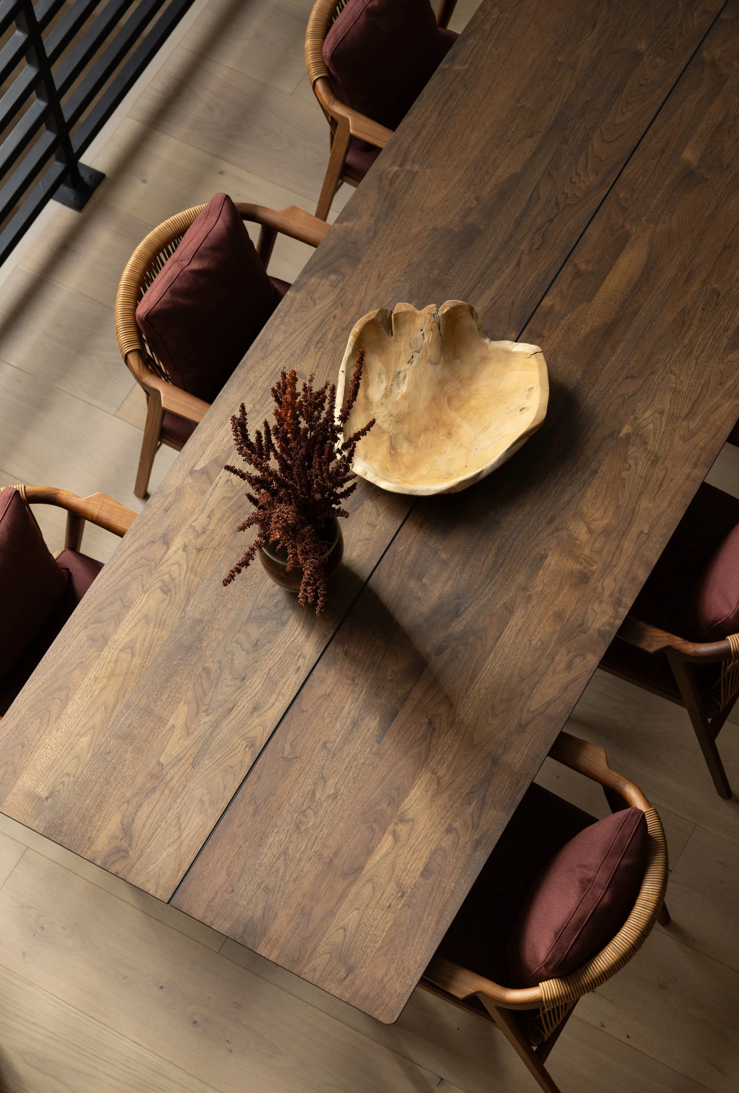 A wooden dining table with a large decorative wooden bowl and a vase of dried flowers in the center, surrounded by six chairs with dark cushions.