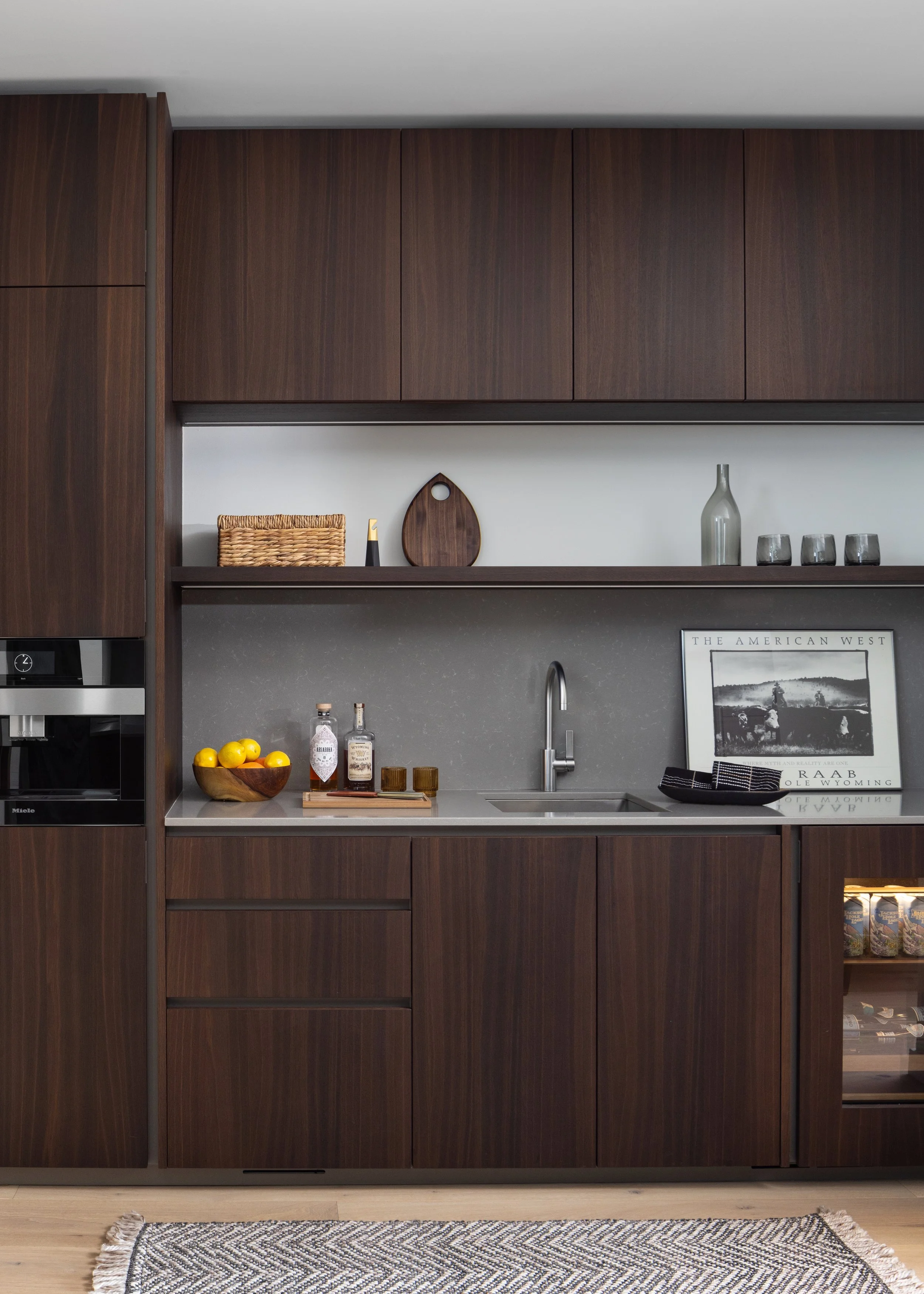 Modern kitchen with dark wood cabinets, gray countertop, stainless steel sink, and minimal decor, including a framed black and white photo and a bowl of lemons.