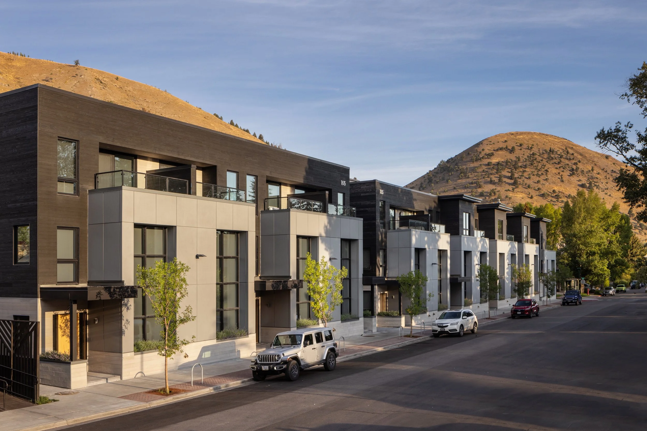 Modern multi-story residential building with balconies, located along a street with parked cars and trees, hills in the background.