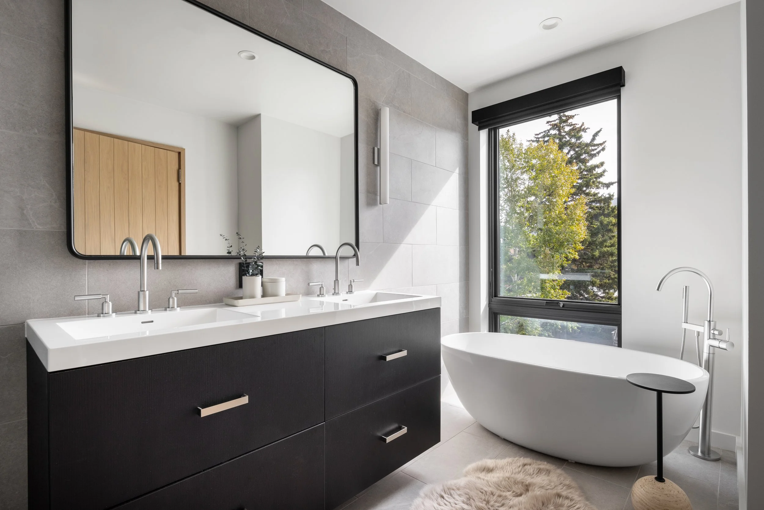 Modern bathroom with a double sink vanity, large mirror, freestanding bathtub, window with a view of trees, and minimal decor.