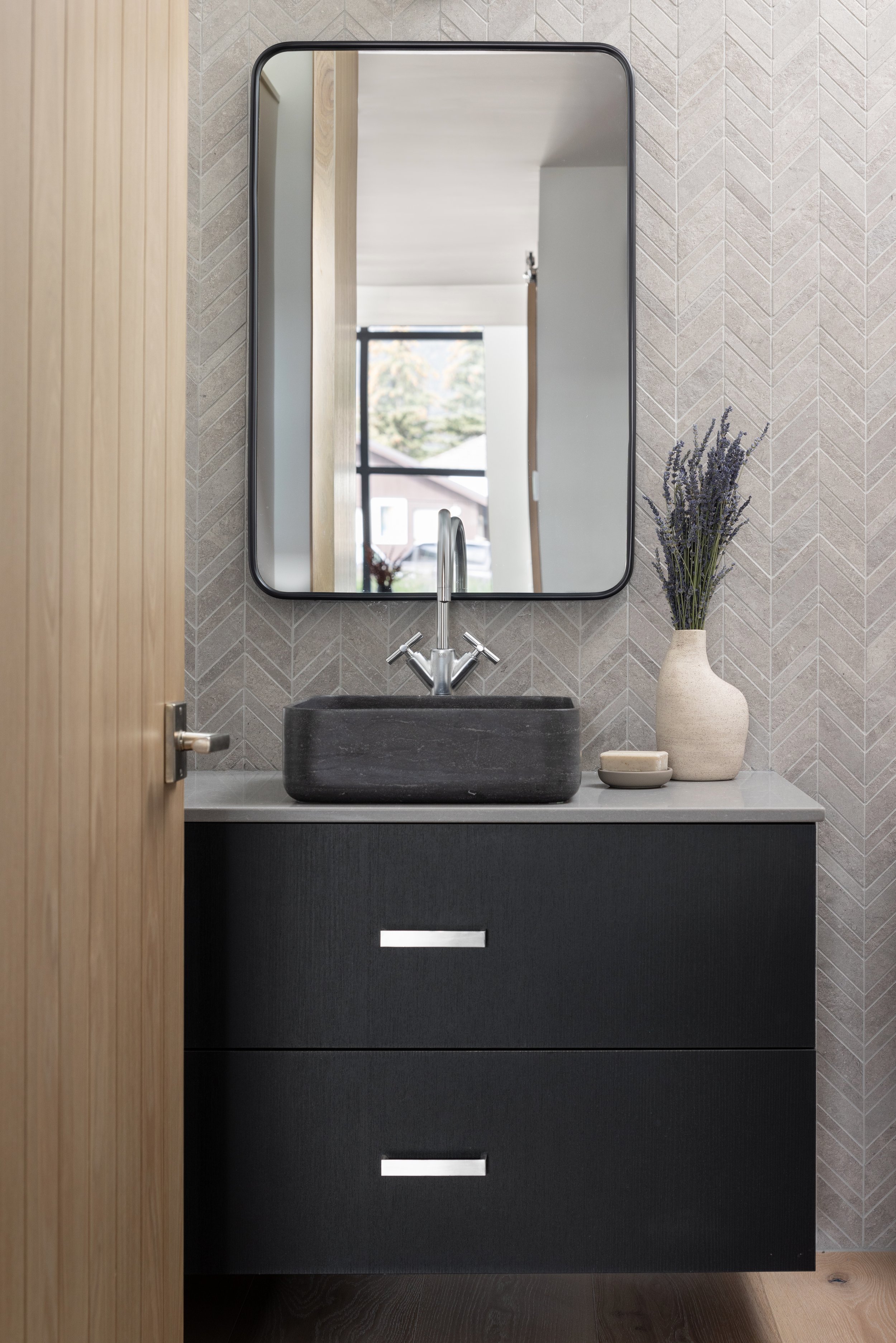 A modern bathroom sink area features a black rectangular stone basin, a sleek chrome faucet, a large mirror above, a minimalist beige vase with lavender flowers, and a small ceramic dish on a light gray countertop. The black vanity has two drawers wi