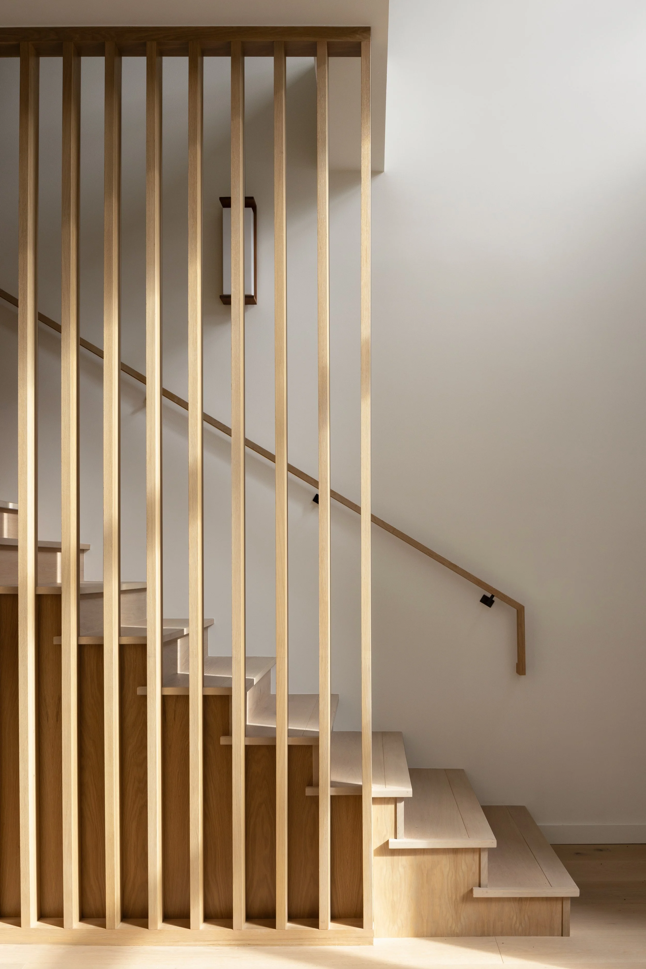 Photo of a wooden staircase with vertical slats on one side, a wooden handrail, and a small wall-mounted light fixture in a modern interior.