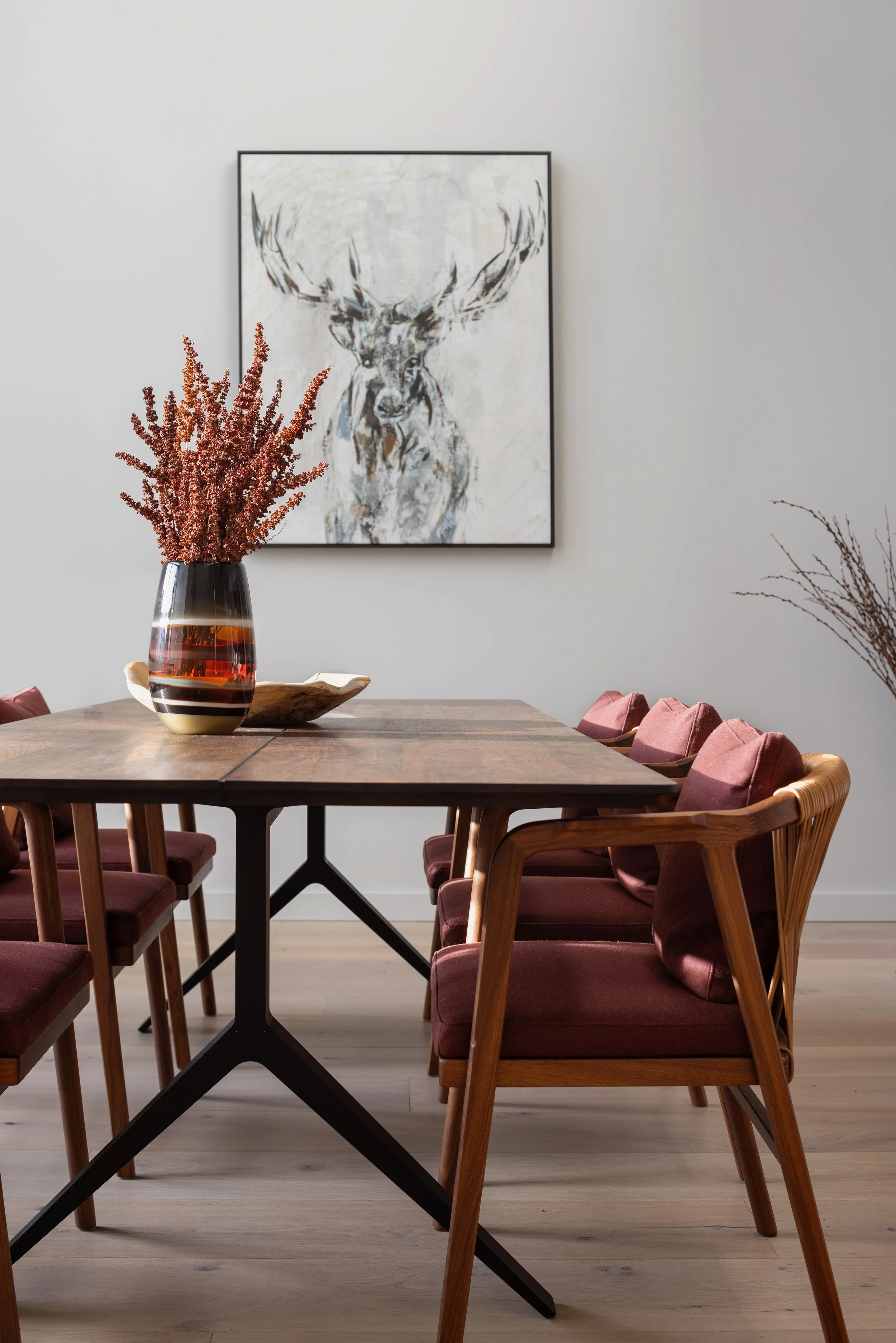 A dining room with a wooden table, six pink-cushioned chairs, a decorative vase with pink flowers, a framed deer painting on the wall, and a light wood floor.