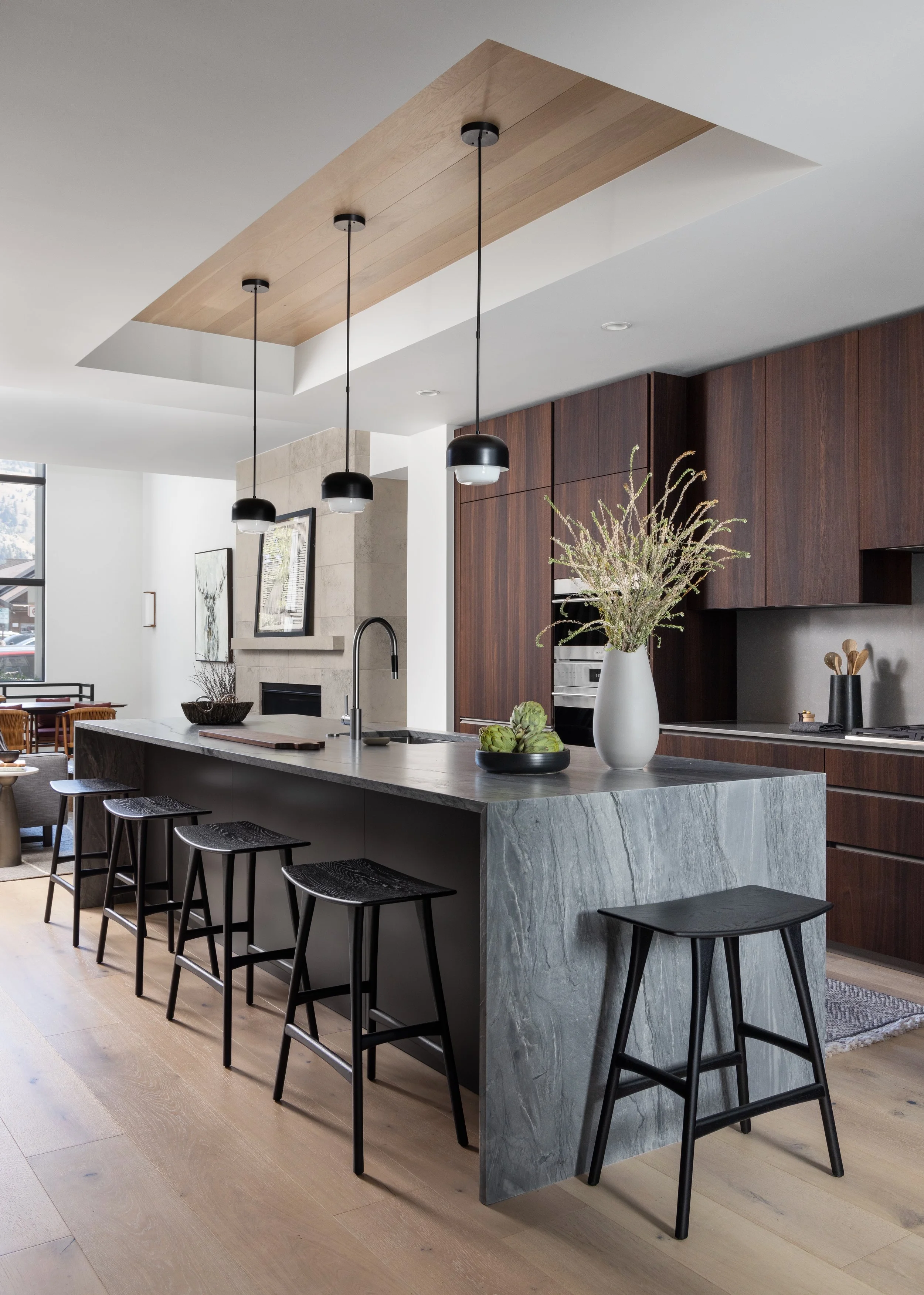 Modern kitchen with gray marble island, black barstools, wooden cabinetry, pendant lights, and decorative plants.