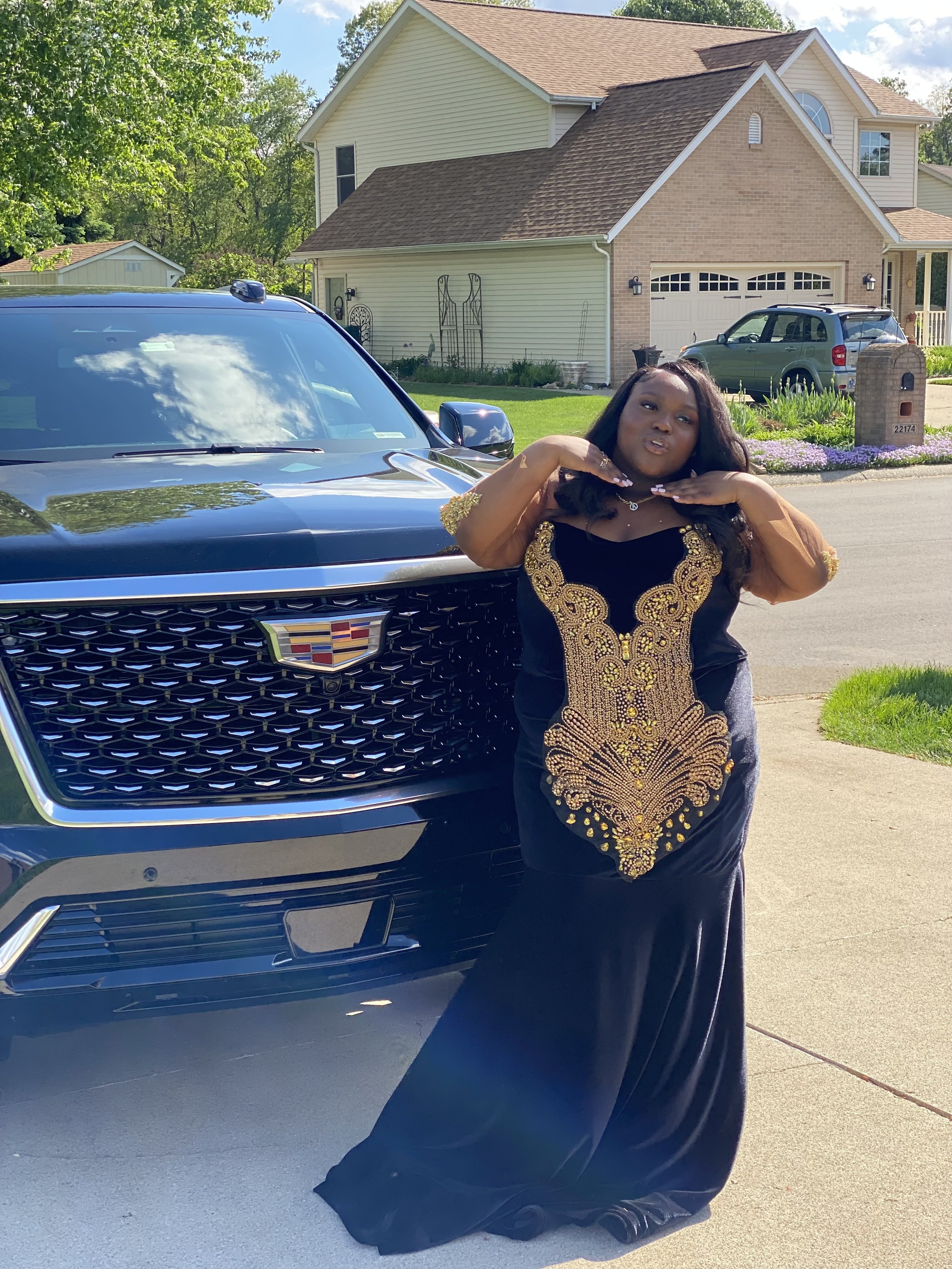 A woman in a black and gold embellished gown standing next to a black Cadillac SUV in a suburban neighborhood, with houses, cars, and trees in the background.