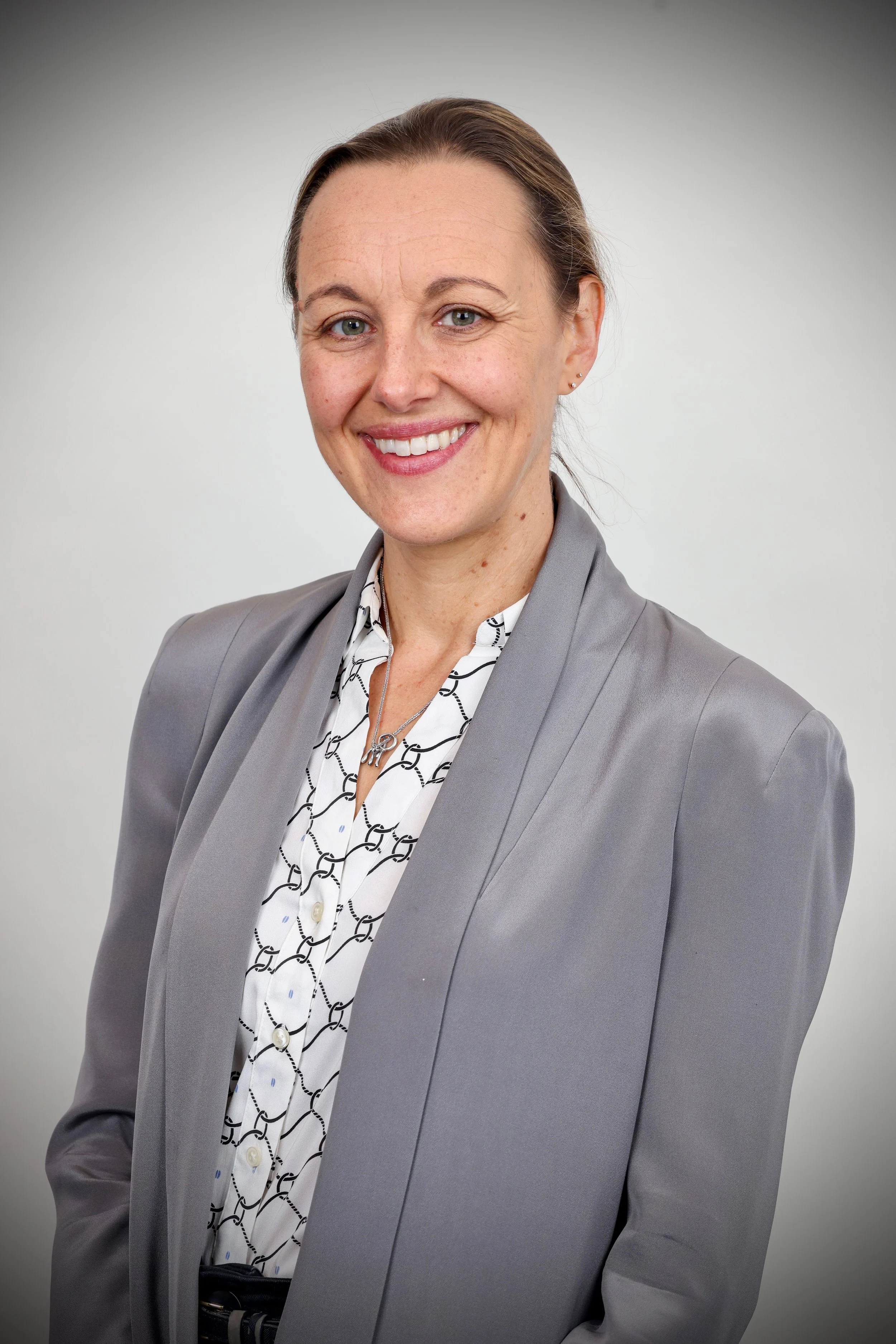 A woman wearing a grey blazer, white patterned shirt, and a silver necklace smiling in front of a plain grey background.