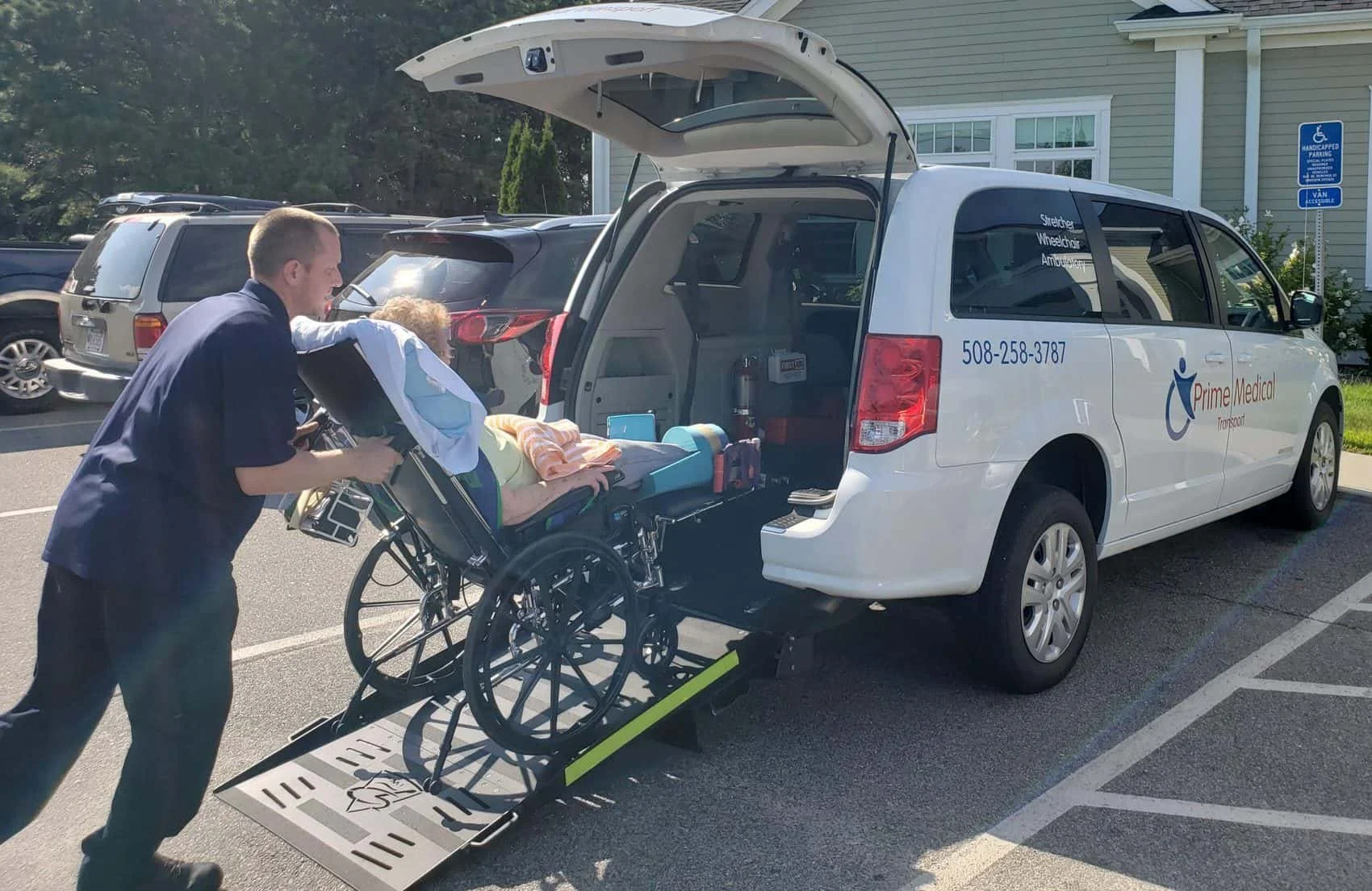 man pushes elderly woman in a stretcher chair into a NEMT van on a sunny day