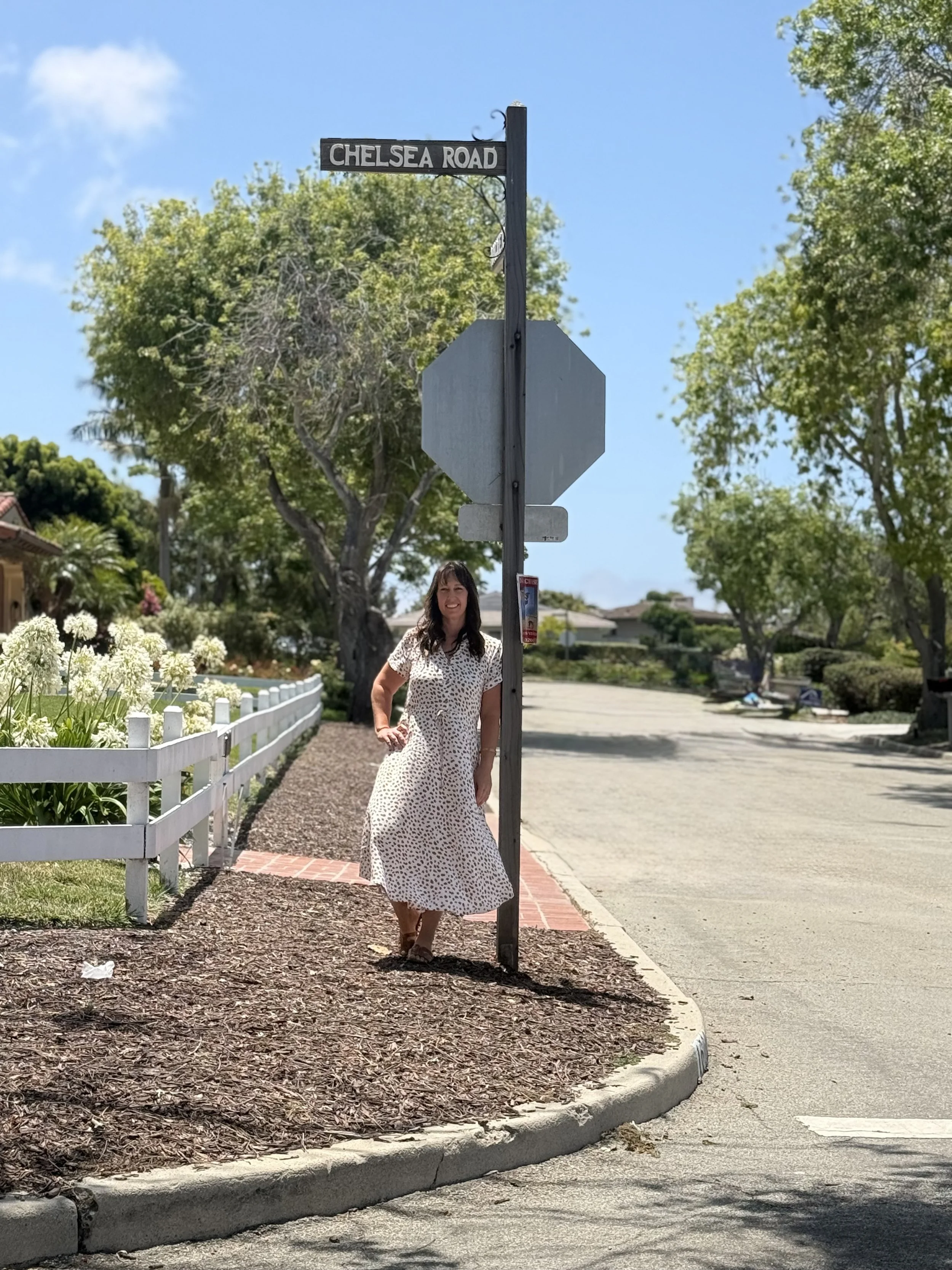 Woman in a white dress with black polka dots standing beside a street sign that reads Chelsea Road. She is smiling and standing on a sidewalk with trees and houses in the background.