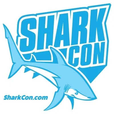 Logo for Shark Con. It features blue sans-serif text and a blue and white illustrated shark. The words "sharkcon.com" are under the main logo image.