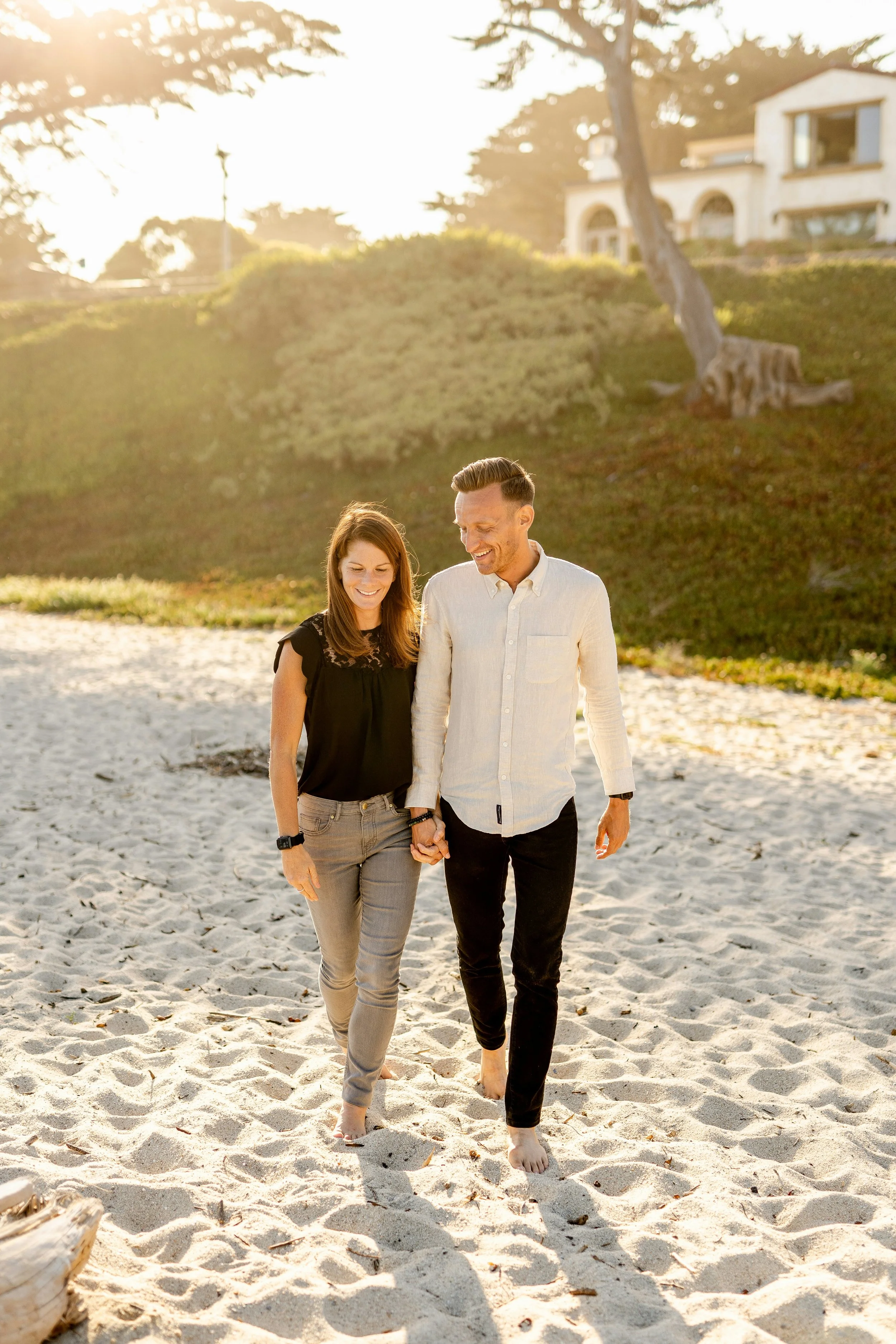 A couple walking barefoot on a sandy beach, holding hands and smiling, with a house and trees in the background during sunset.
