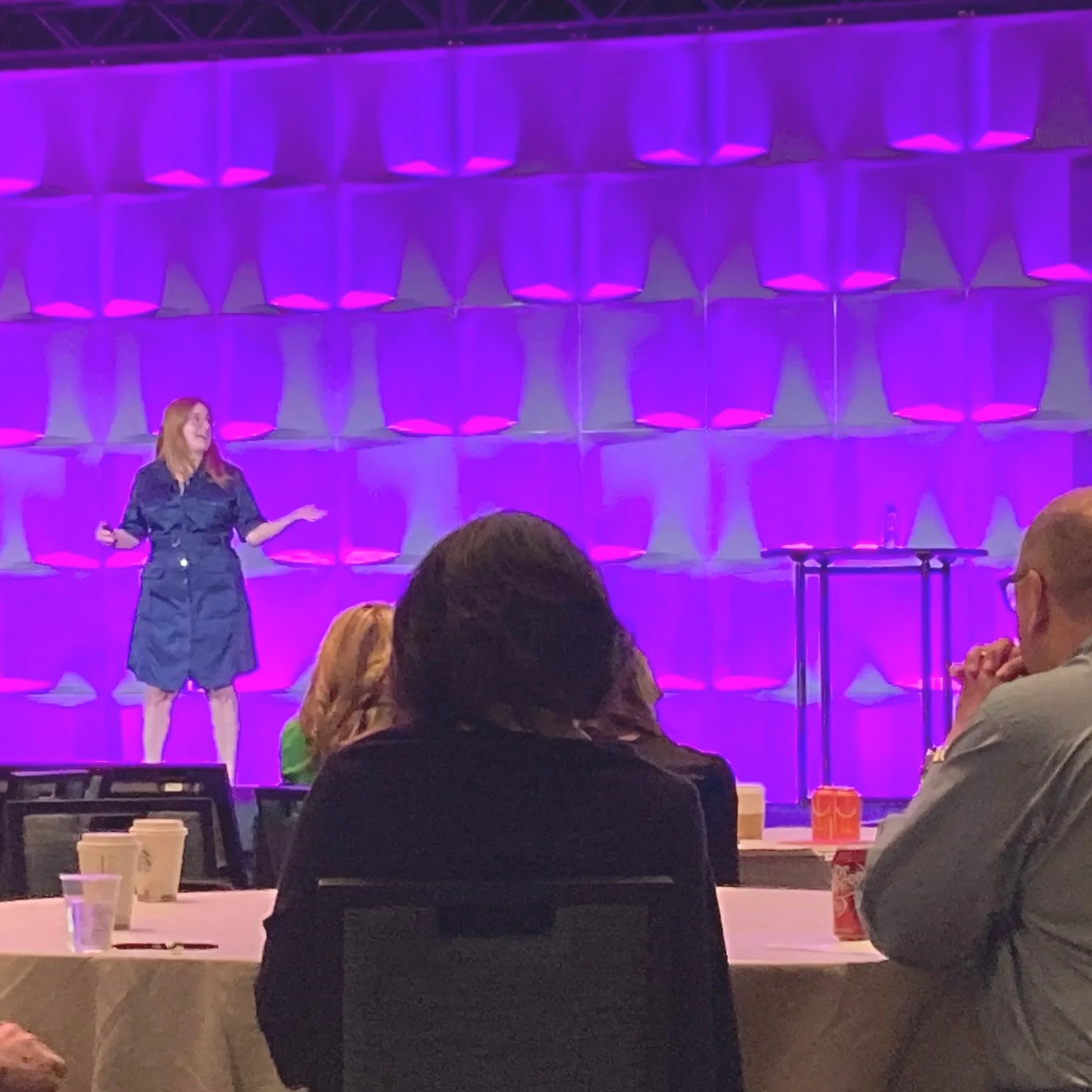 A woman in a black dress presenting on stage with purple lighting. Several audience members seated at tables in front, some with drinks and coffee cups, watching her perform.