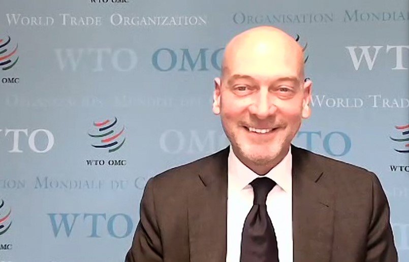 Man with a bald head and beard, smiling, wearing a dark suit, white shirt, and black tie, standing in front of a blue background with WTO and World Trade Organization logos.