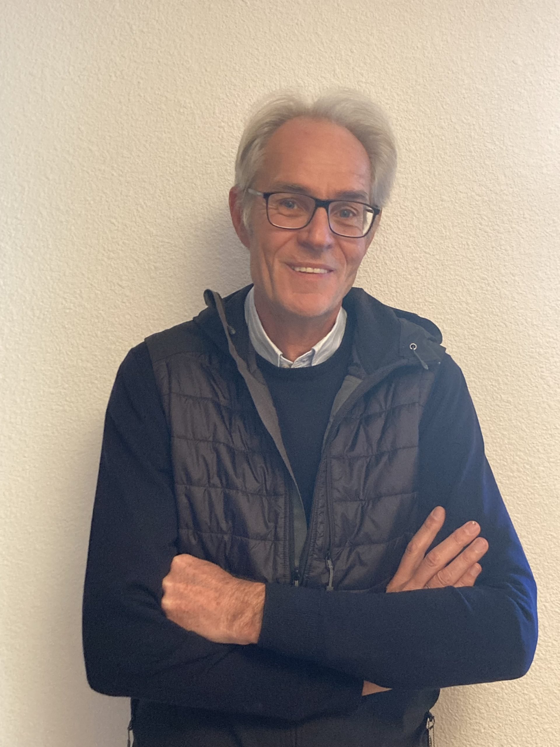 A middle-aged man with gray hair and glasses, wearing a black jacket over a white shirt, standing in front of a plain beige wall, smiling with arms crossed.