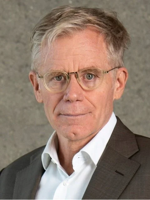 A middle-aged man with light-colored hair, glasses, wearing a white shirt and a dark suit, against a gray background.