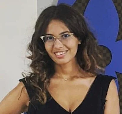 A woman with curly dark hair wearing glasses and a black sleeveless top, smiling in front of a geometric patterned background.