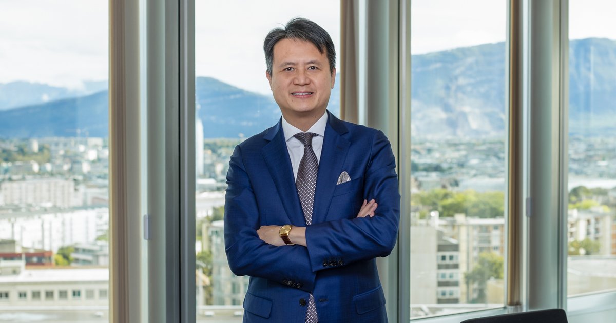 A man in a blue suit with a white shirt and patterned tie standing with crossed arms in front of large windows overlooking a cityscape and mountains.