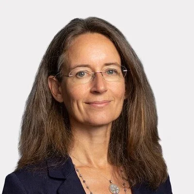 A professional woman with long brown hair, wearing glasses and a dark blazer, smiling slightly against a plain white background.