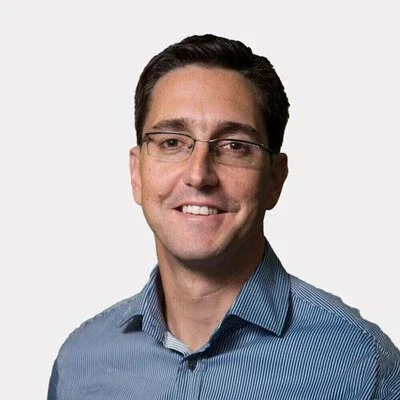 A man with short dark hair, glasses, and a light blue dress shirt, smiling against a light background.