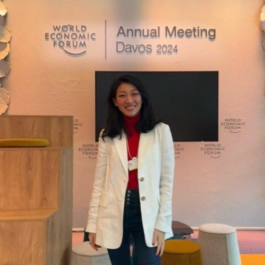 A woman standing in front of a backdrop at the World Economic Forum Annual Meeting in Davos 2024, smiling and wearing a white blazer over a red top.