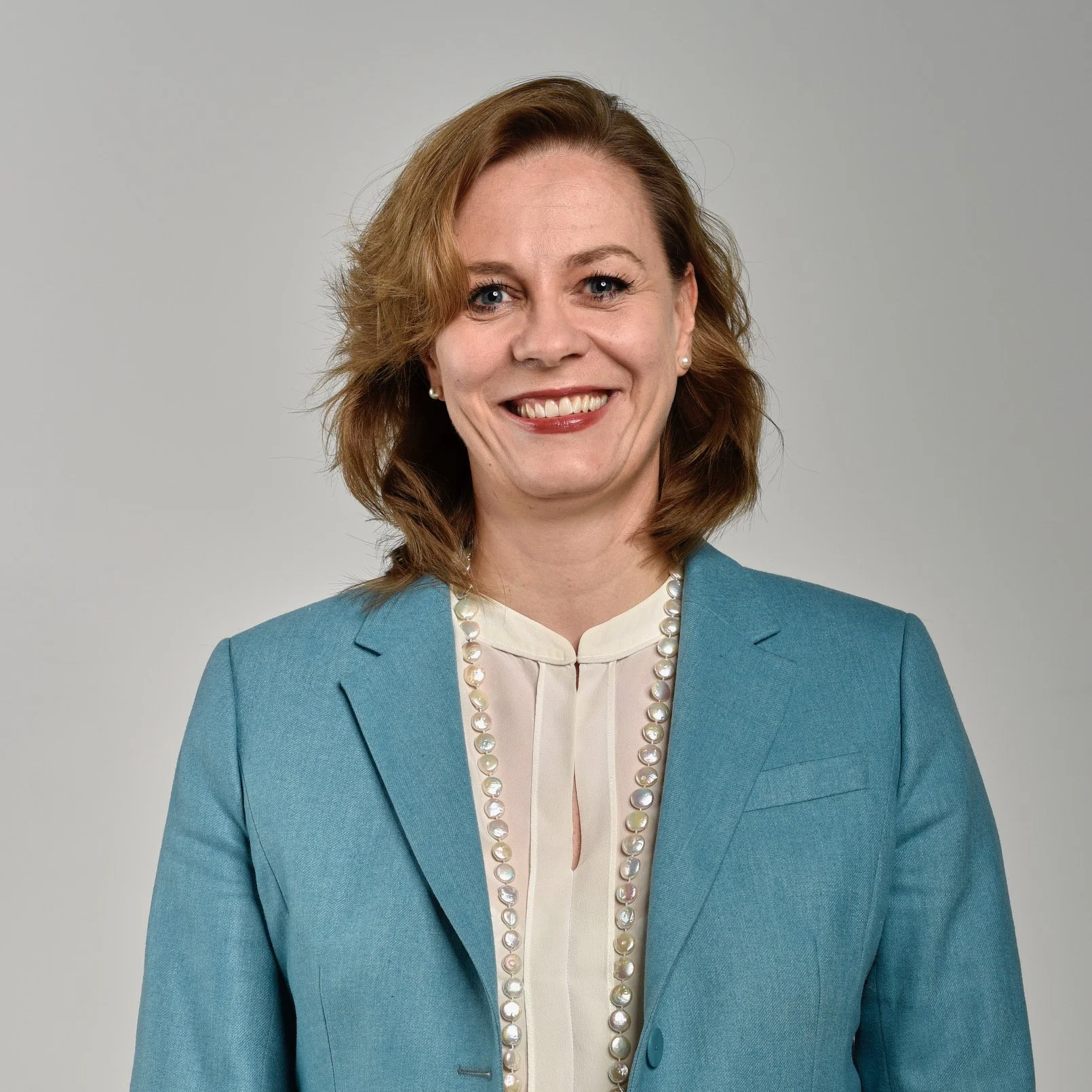 A professional woman with shoulder-length brown hair, wearing a light blue blazer, cream blouse, and a pearl necklace, smiling at the camera against a plain neutral background.