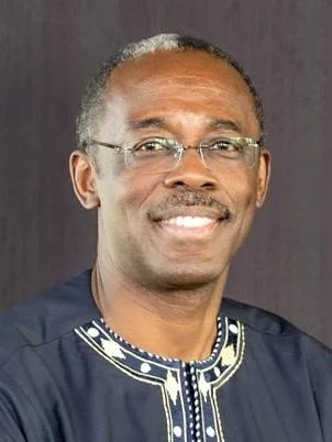 Portrait of a smiling man with glasses and a traditional embroidered shirt, against a dark background.