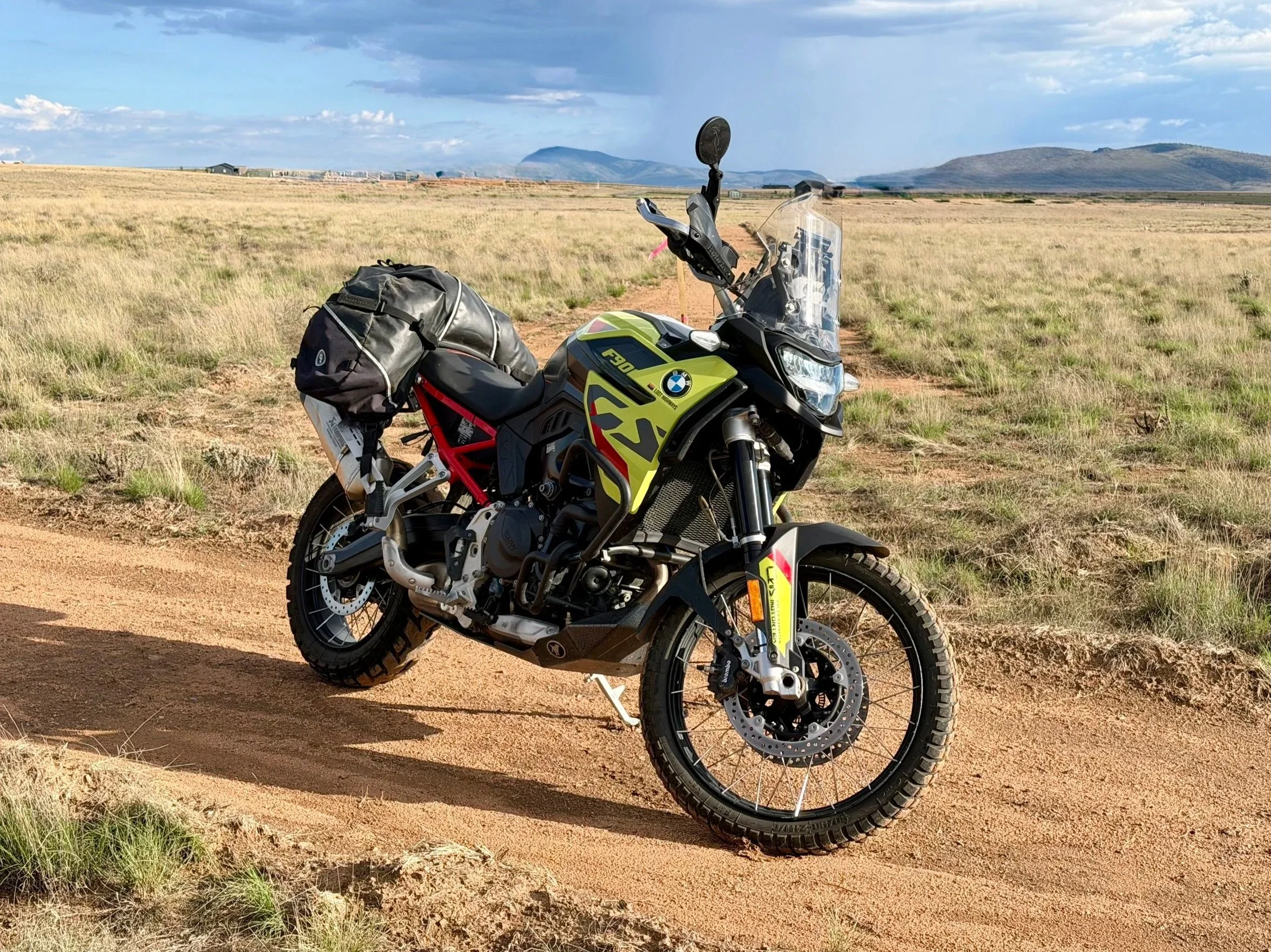 A BMW adventure motorcycle parked on a dirt trail in an outdoor setting with greenery and trees in the background.