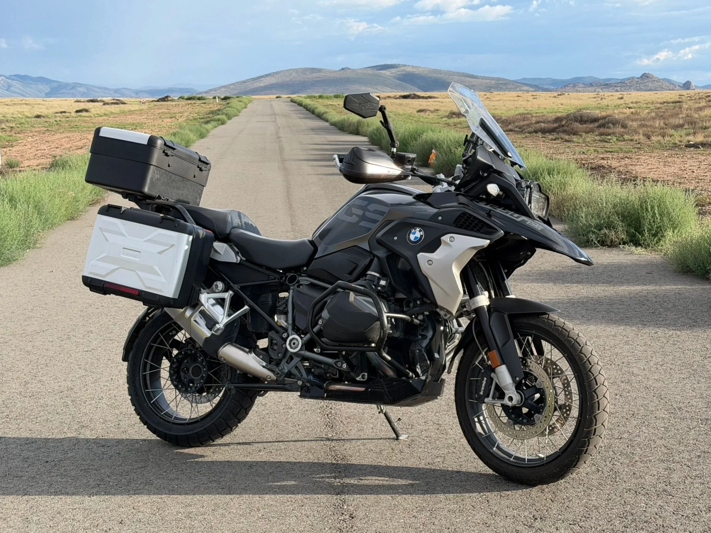 Black BMW adventure motorcycle with side and top luggage cases parked on a dirt road with mountains and a sunset sky in the background.