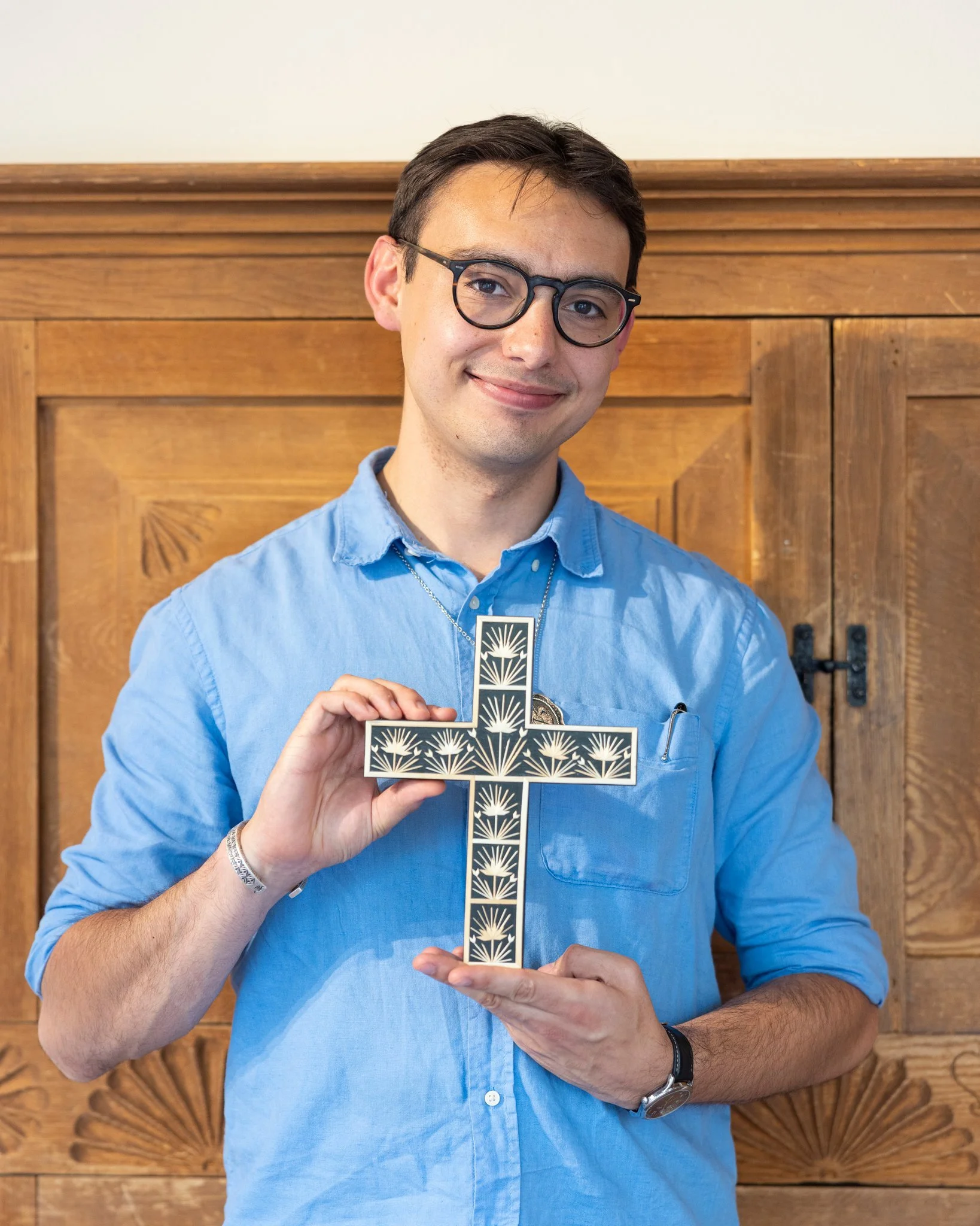 A man with short brown hair and glasses weaing a blue button down shirt holds a black cross decorated with straw designs.
