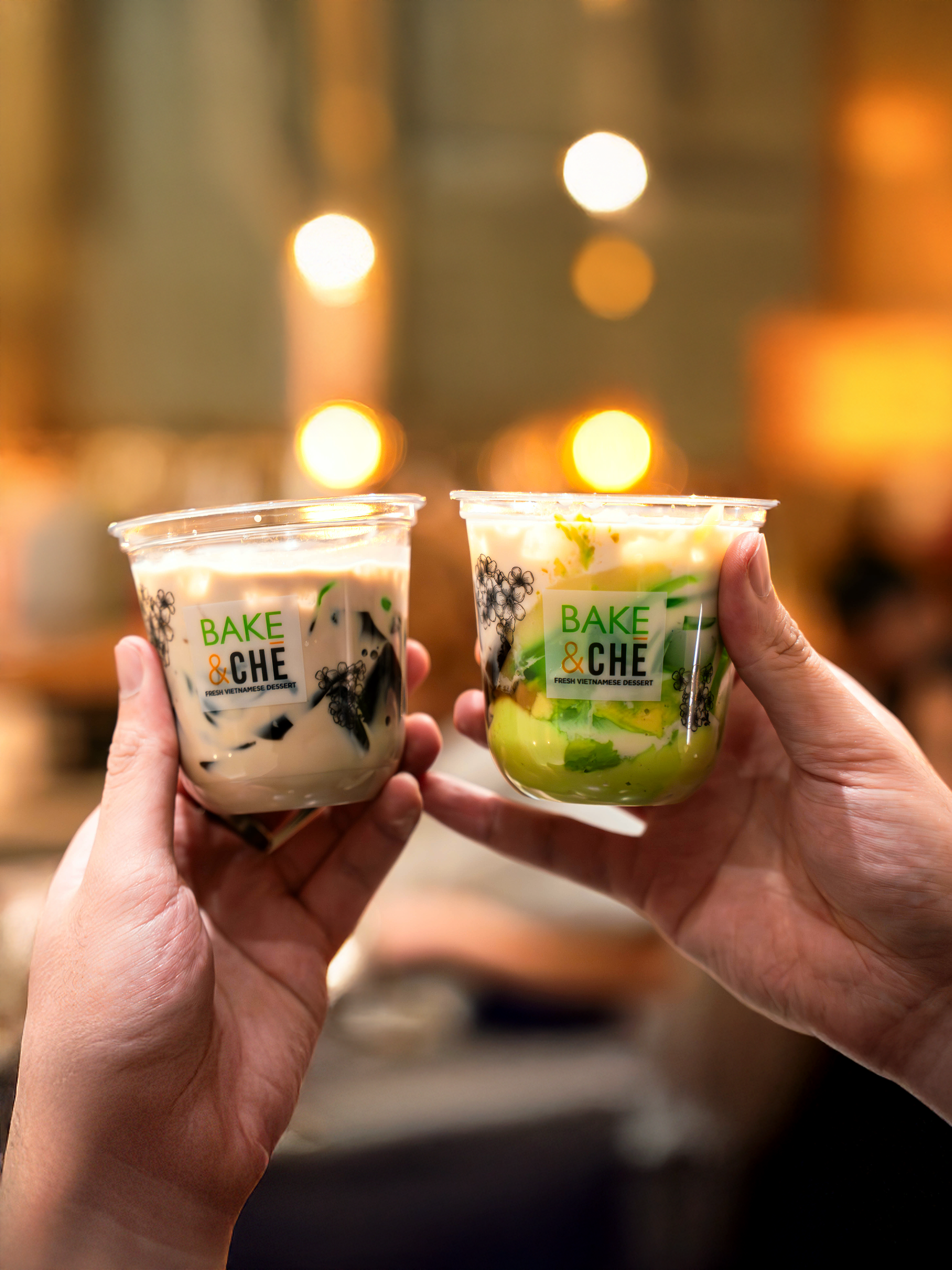 Two hands holding plastic cups with desserts and the label "Bake & Che", one with black jelly and the other with green jelly, in a blurred indoor setting with warm lights.