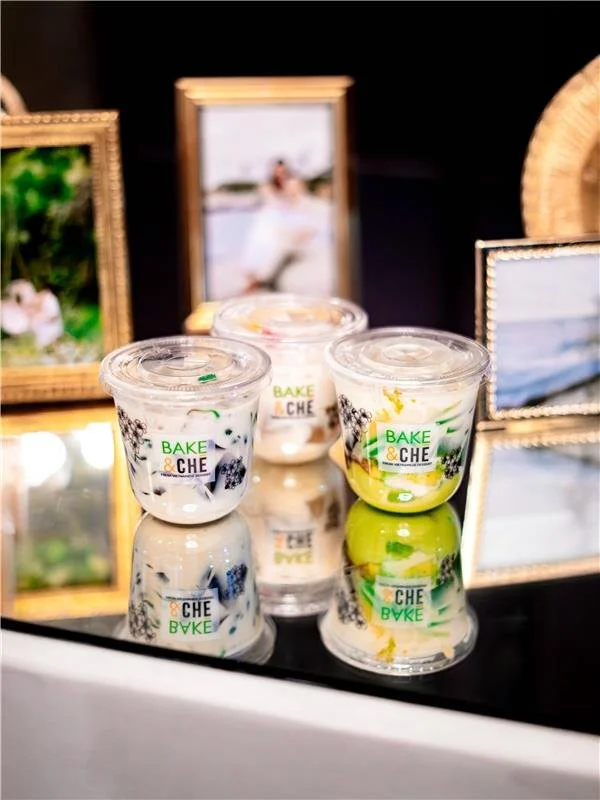 Three small jars with plastic lids labeled 'Bake & Che', placed on a reflective surface with framed photographs in the background.