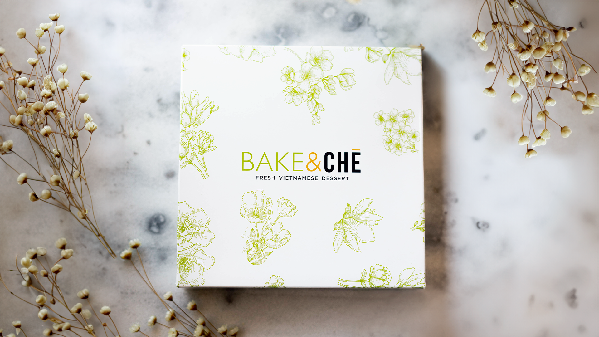 A white box with green floral illustrations and the text "BAKE & CHÈ" in green, orange, and black, placed on a marble surface with dried white flowers surrounding it.