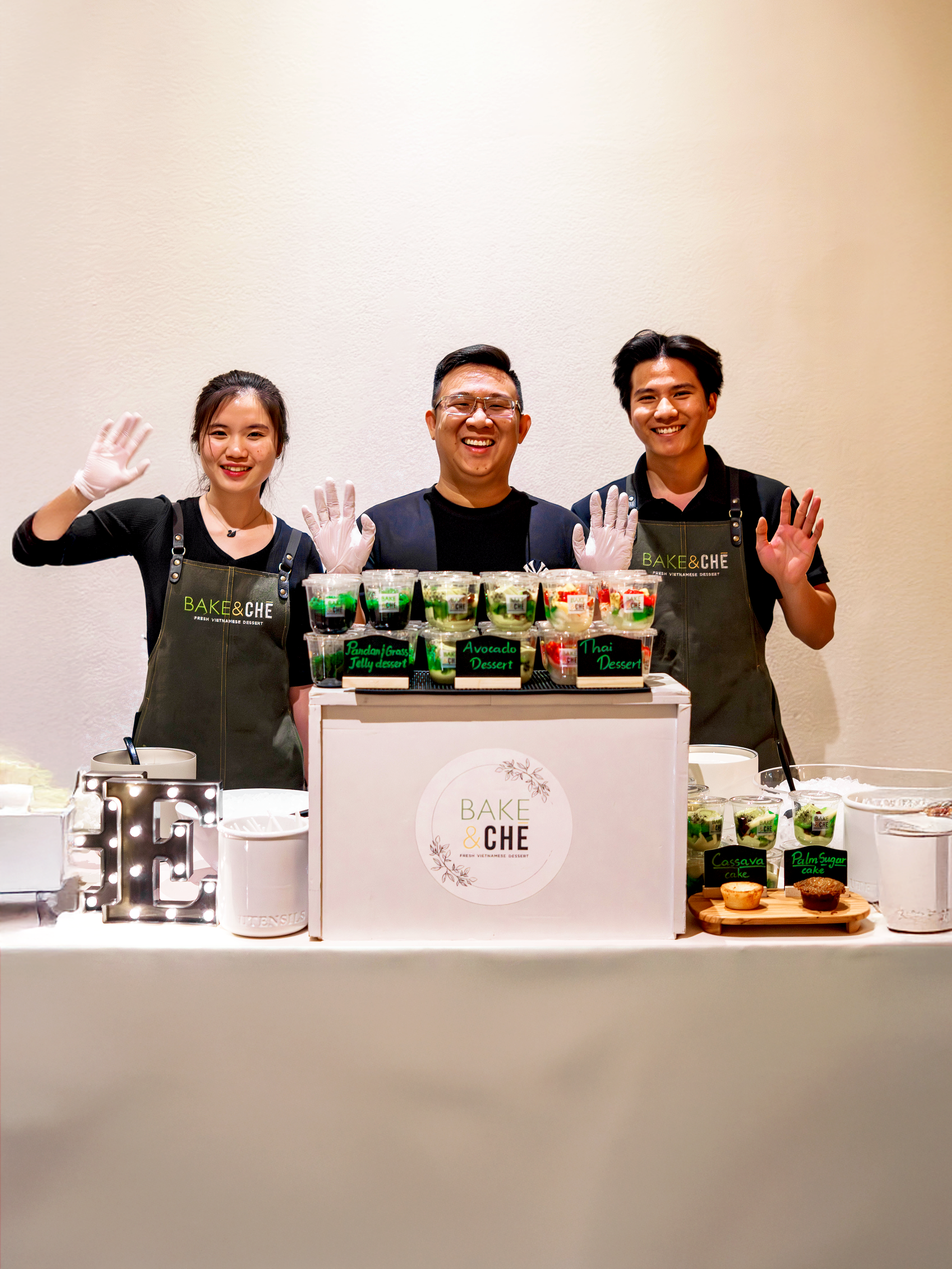 Three people standing behind a dessert stall, smiling and waving, with various colorful desserts on display.