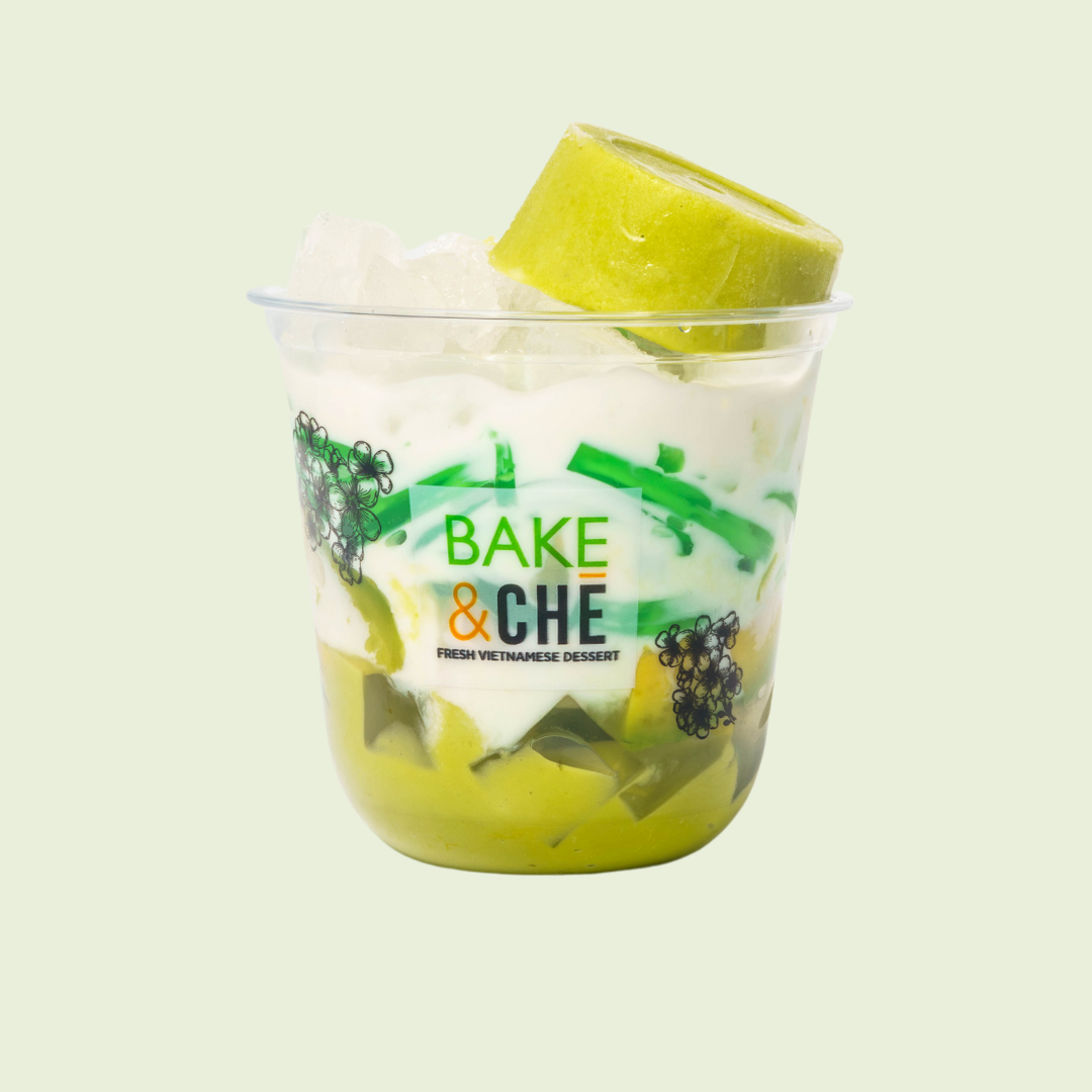 A clear plastic cup filled with a Vietnamese dessert featuring green jelly, white creamy layers, and a piece of green fruit on top, with a label reading 'Bake & Che, Fresh Vietnamese Dessert.'