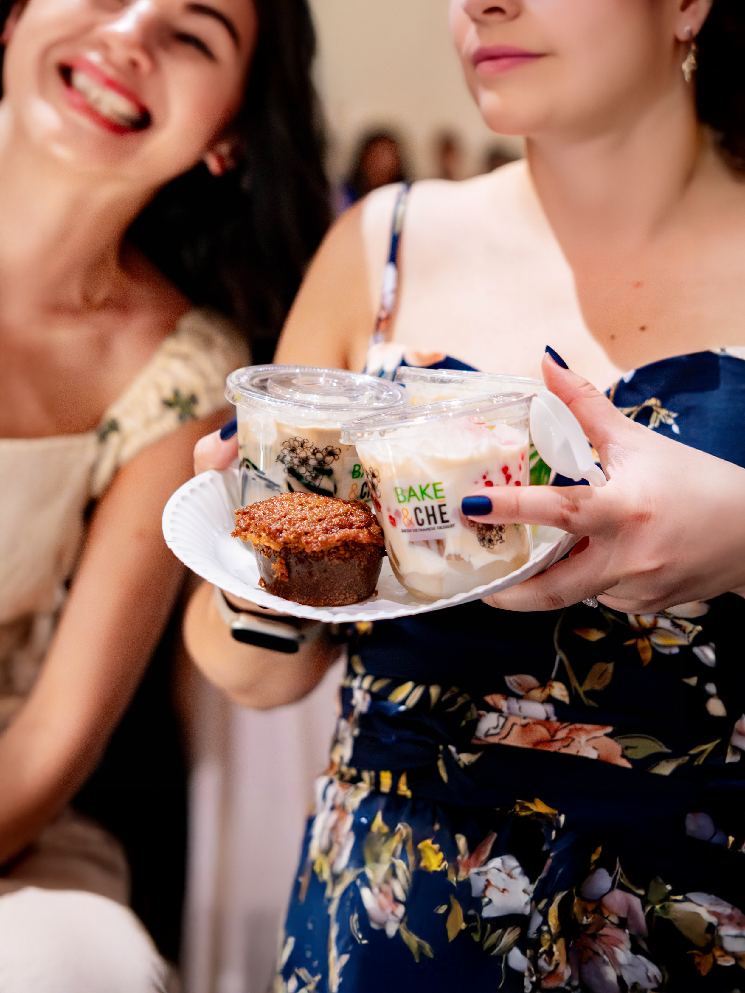 A woman is holding a tray with four small dessert cups and a muffin, while smiling with another woman next to her, at a social gathering or event.