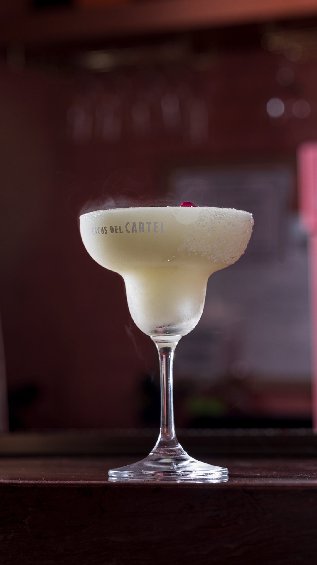 Two yellow cocktails with salted rims served in margarita glasses on an outdoor table, with a background of green plants and a partly cloudy sky.