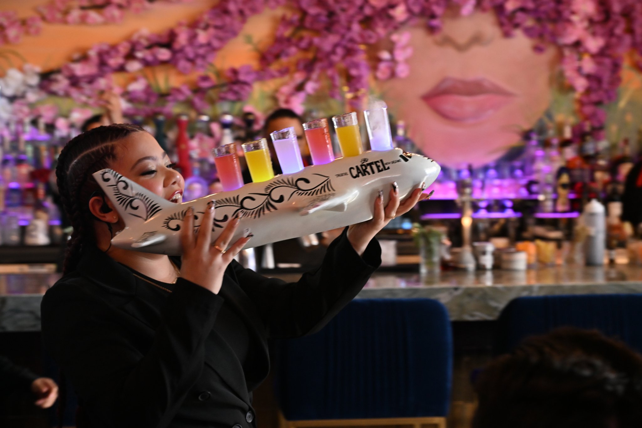 A woman serving cocktails from a tray shaped like an airplane at a bar with pink floral artwork and a large face painting in the background.