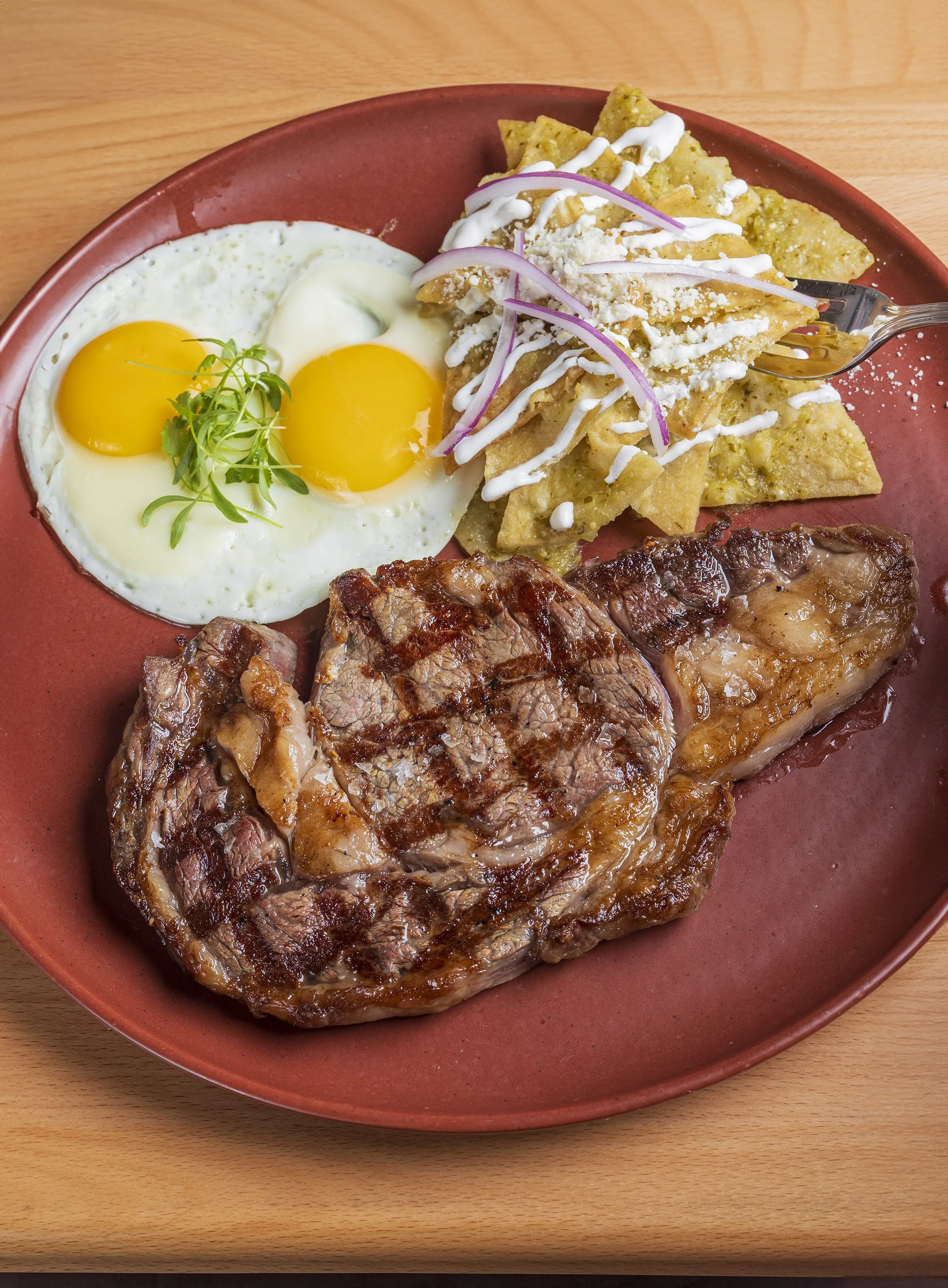 Plate with sunny-side-up eggs, grilled steak, and a side of potato chips with onions and drizzled sauce.