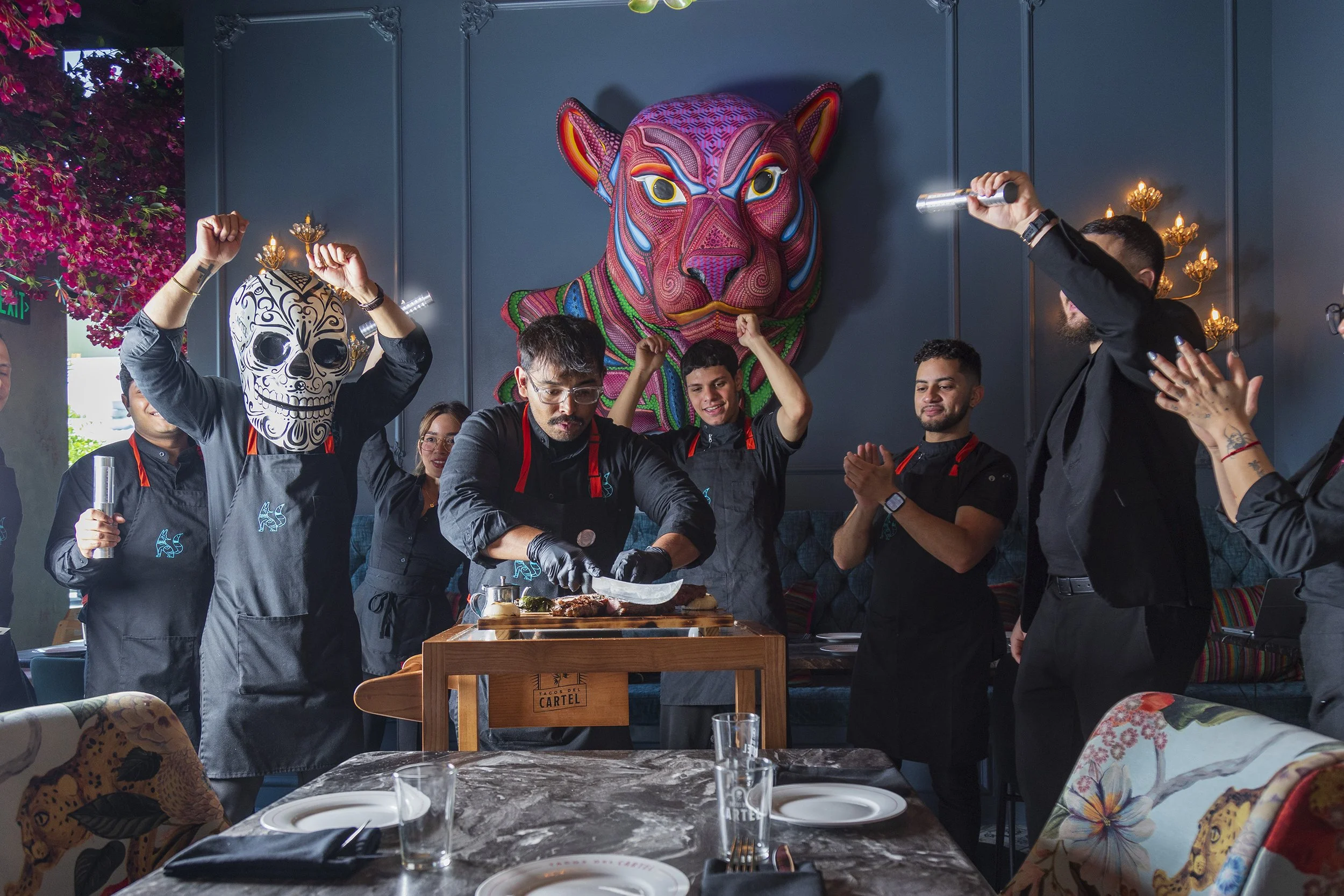 Group of restaurant staff celebrating with some wearing masks, some with arms raised in victory in a decorated dining area with colorful wall art