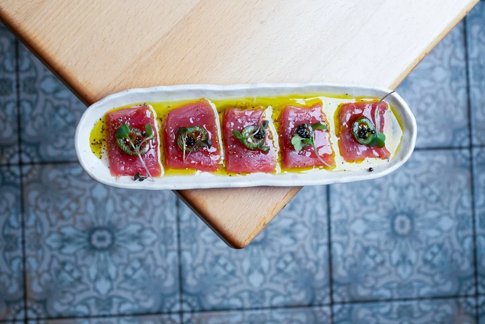 A white oval dish with five slices of raw tuna topped with microgreens, black caviar, and drizzled with olive oil, on a wooden table.