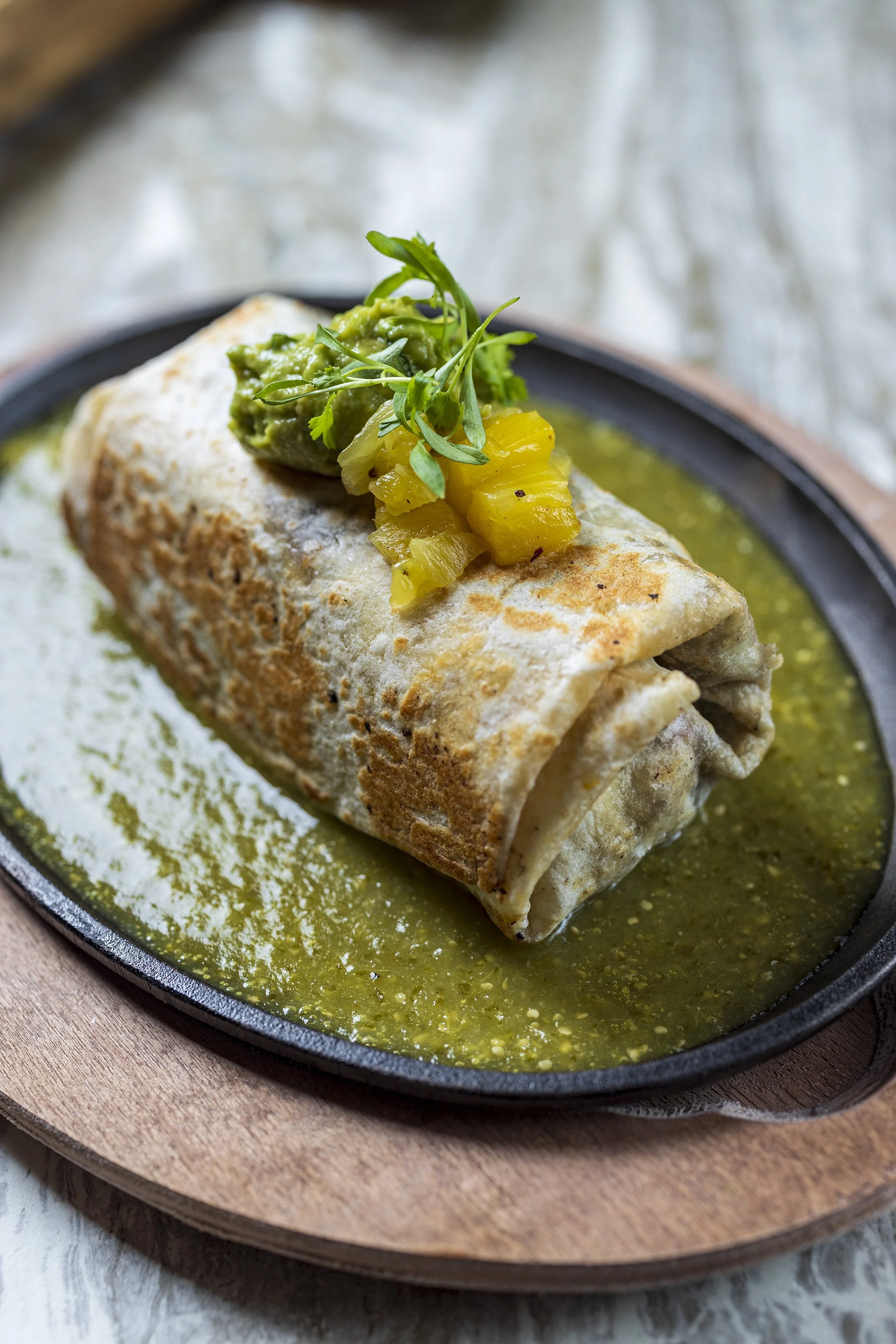 A rolled burrito with a crispy exterior on a black skillet, topped with guacamole, diced yellow pickles, and microgreens, served with green sauce around it.