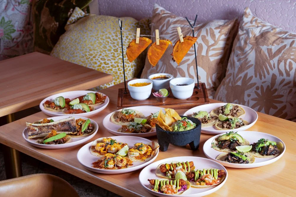 A table with various Mexican foods including tacos, nachos, and salsas, with a background of pillows and beds.