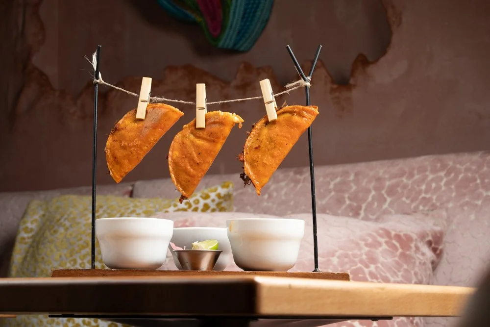 Three pieces of fried food hanging on a string with clothespins, served with a small cup of sauce and two bowls underneath, on a wooden tray.
