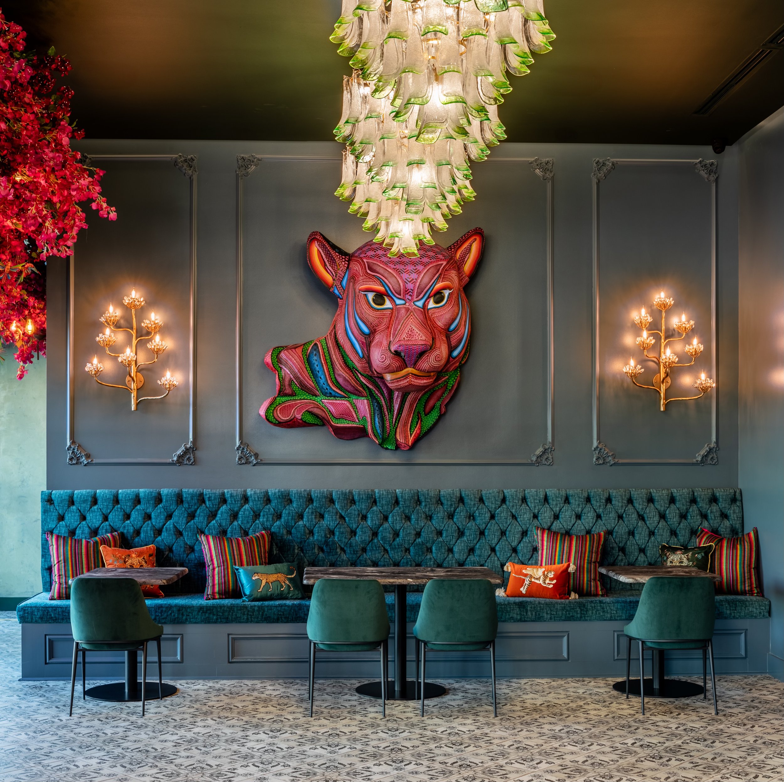 Upscale lounge area with a teal tufted banquette, colorful striped pillows with animal illustrations, and a patterned rug. On the dark wall behind, a large multicolored tiger head sculpture and decorative gold wall sconces. Green glass chandelier han