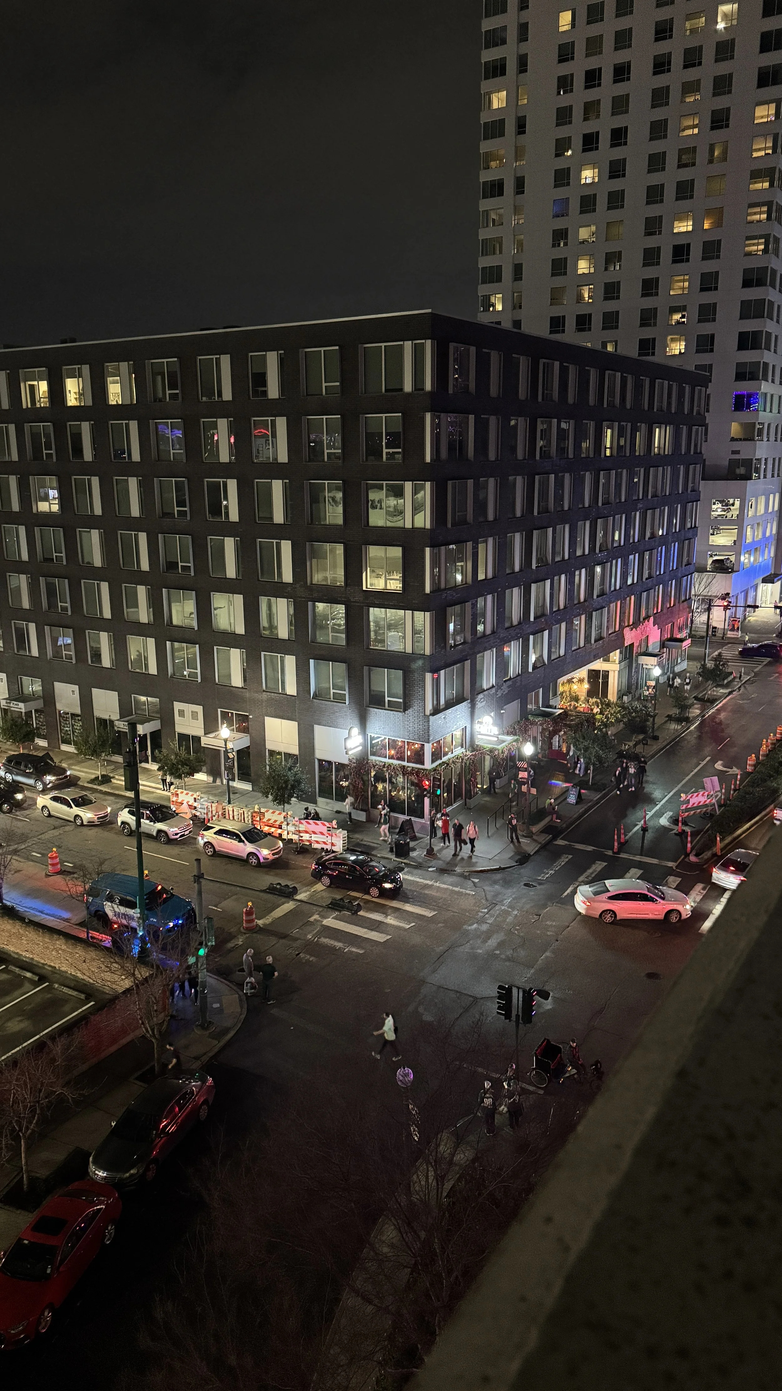 Nighttime city street view showing a multi-story building with lit windows, cars parked along the street, and pedestrians walking on the sidewalk.