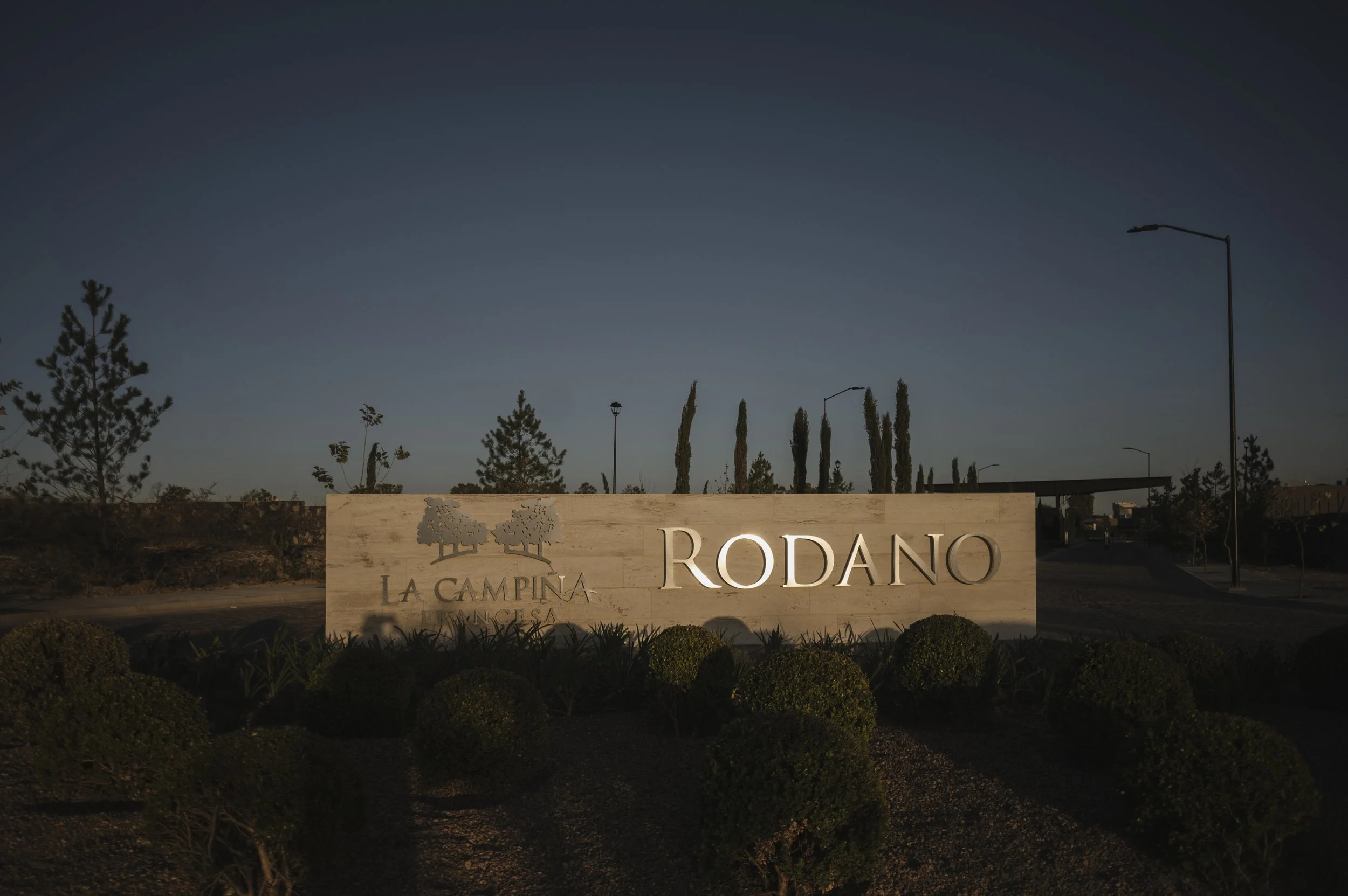 Entrada a la comunidad de Rodano, en La Campina, con cartel de bienvenida y plantas ornamentales en primer plano.