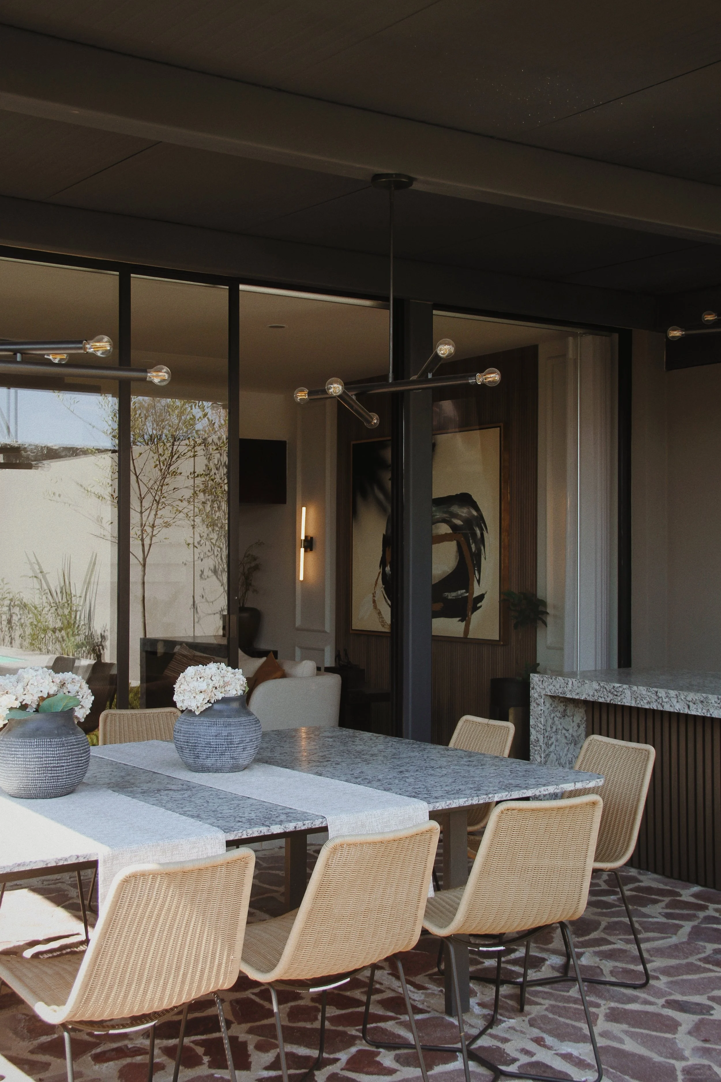 Mesa de comedor con cuatro sillas y jarrones con flores en un espacio interior con ventanas grandes y decoración moderna.